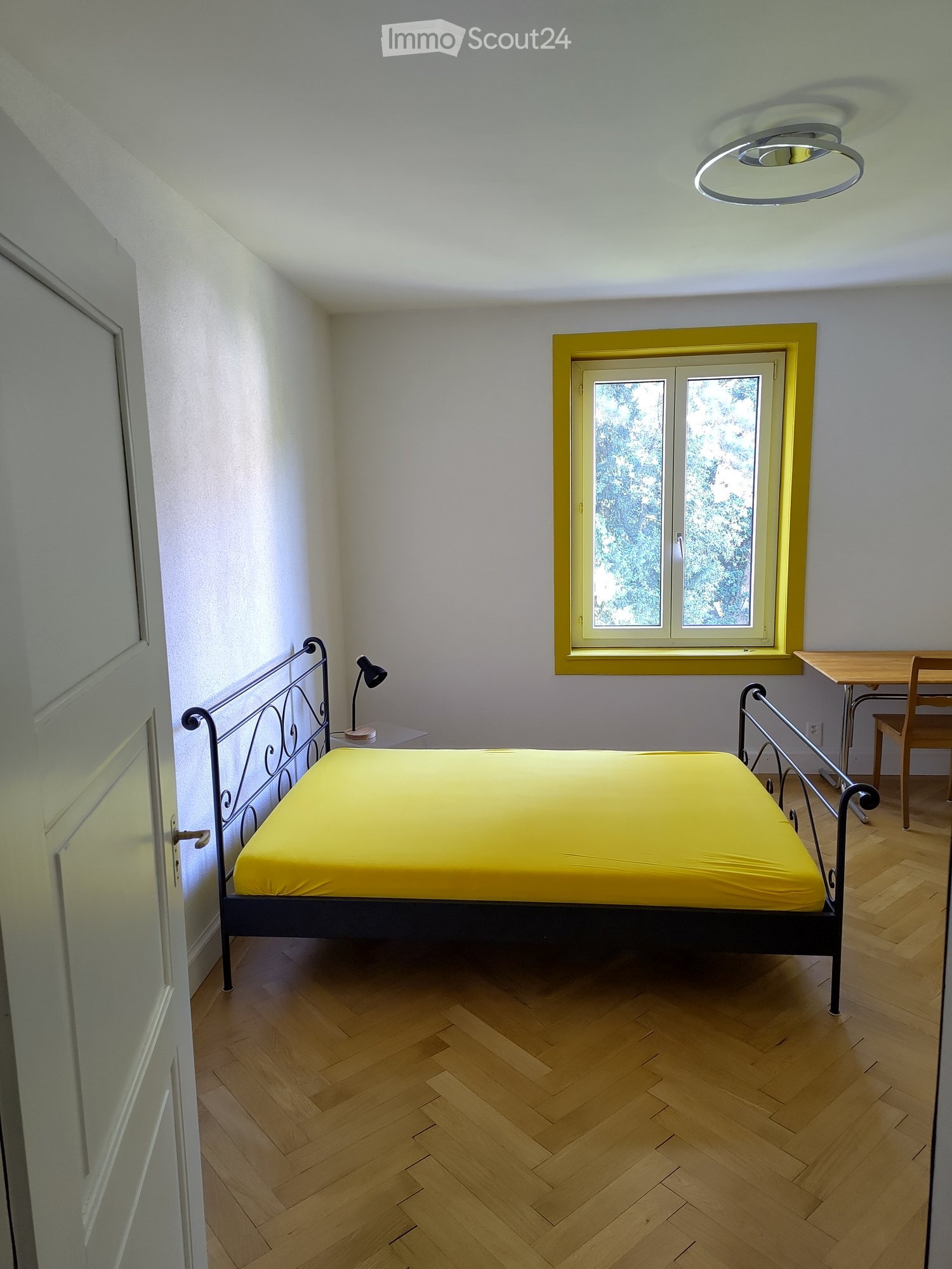 A small bedroom with a wooden floor, a single bed with a yellow bedspread, a small table with a lamp, and a chair near the window.