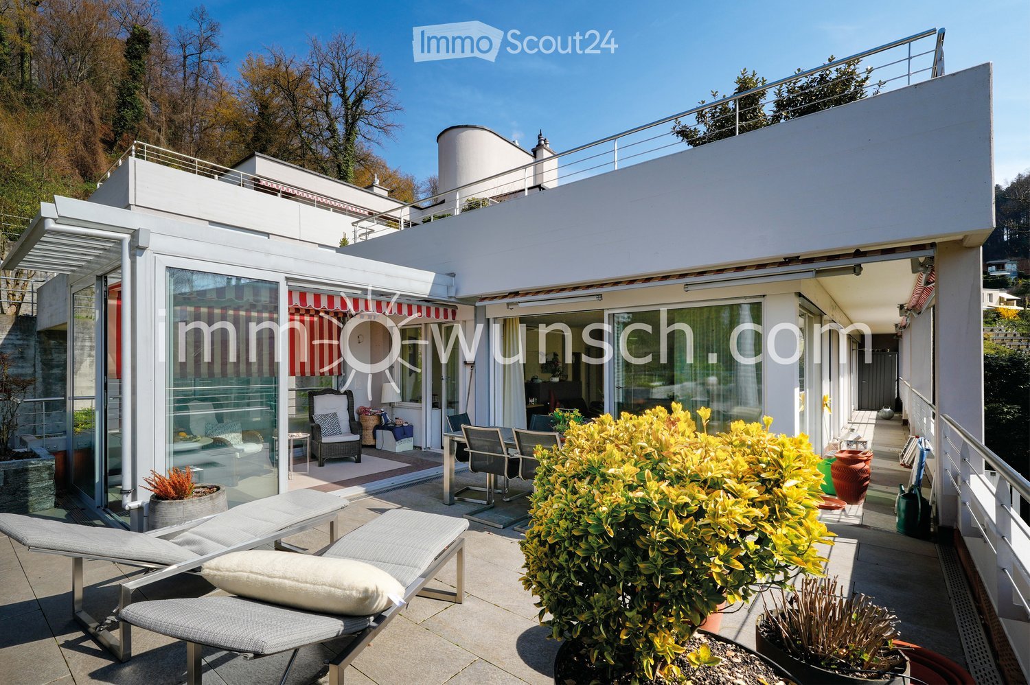 A modern rooftop terrace with lounge chairs, a dining table, potted plants, and a view of the surrounding greenery.