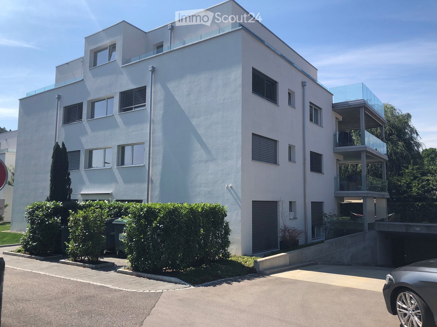 white building, 3 levels, windows with white frames, 2 garages, shrubs and trees in front, entrance on left side