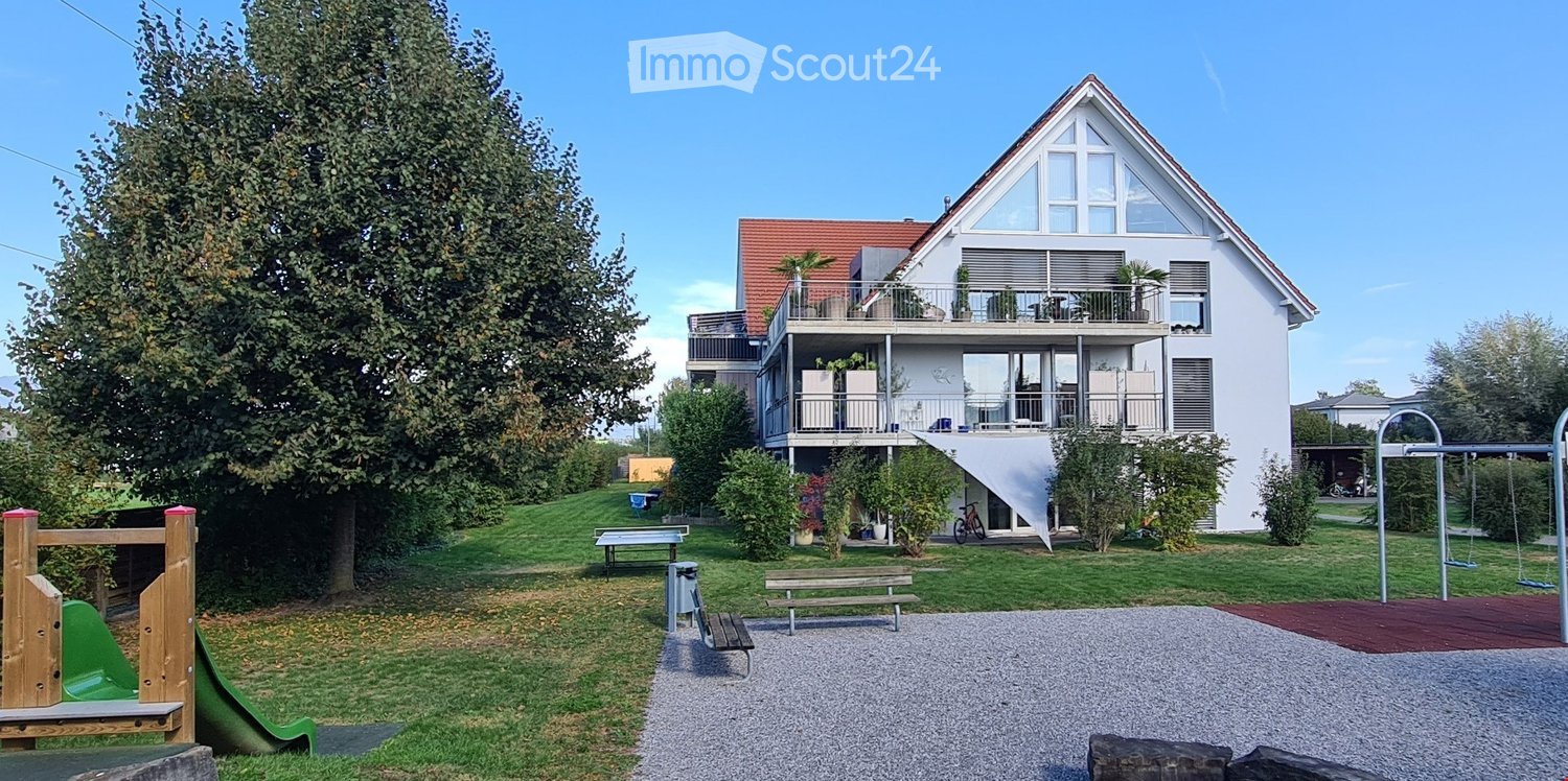 House. A two-story house with a red roof and white walls, featuring a balcony with potted plants and a white awning. The house is surrounded by a well-maintained garden with various plants, trees, and a gravel pathway. There is a playground with a slide,