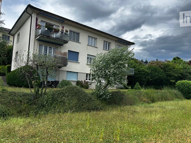 Multi-story residential building with balconies, surrounded by greenery and a grassy area.