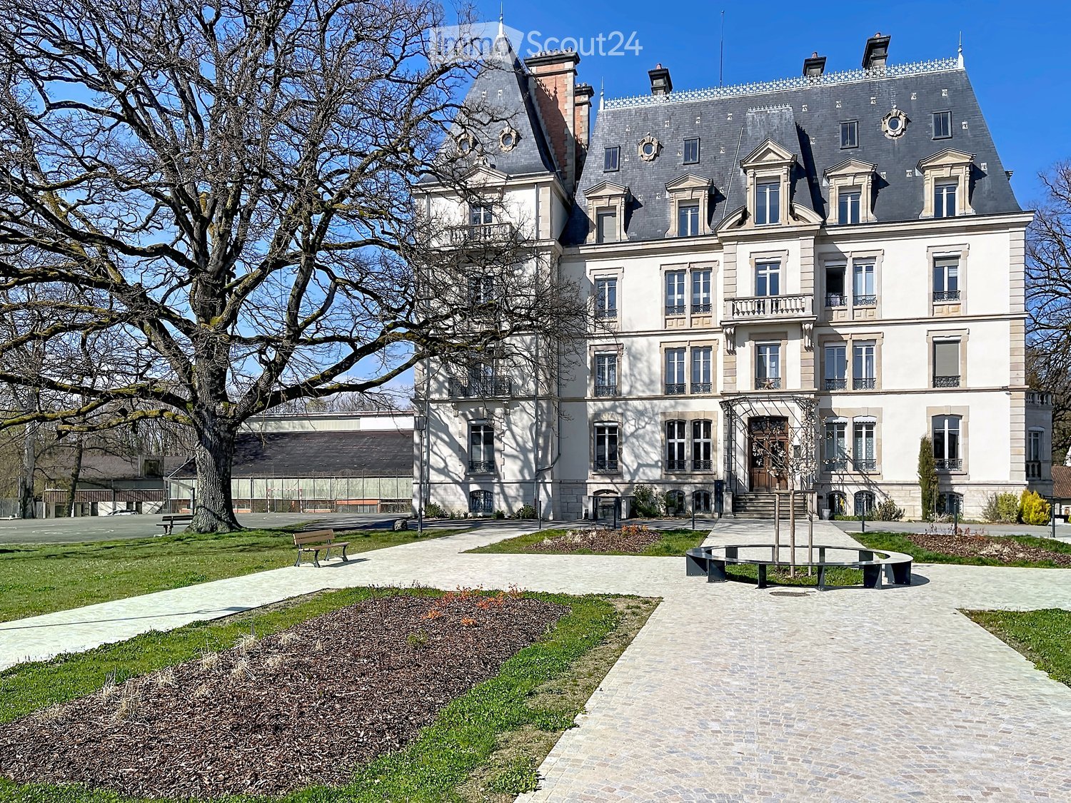Large, elegant mansion with multiple stories, grey roof, white walls, numerous windows, and a spacious garden area with pathways and benches.