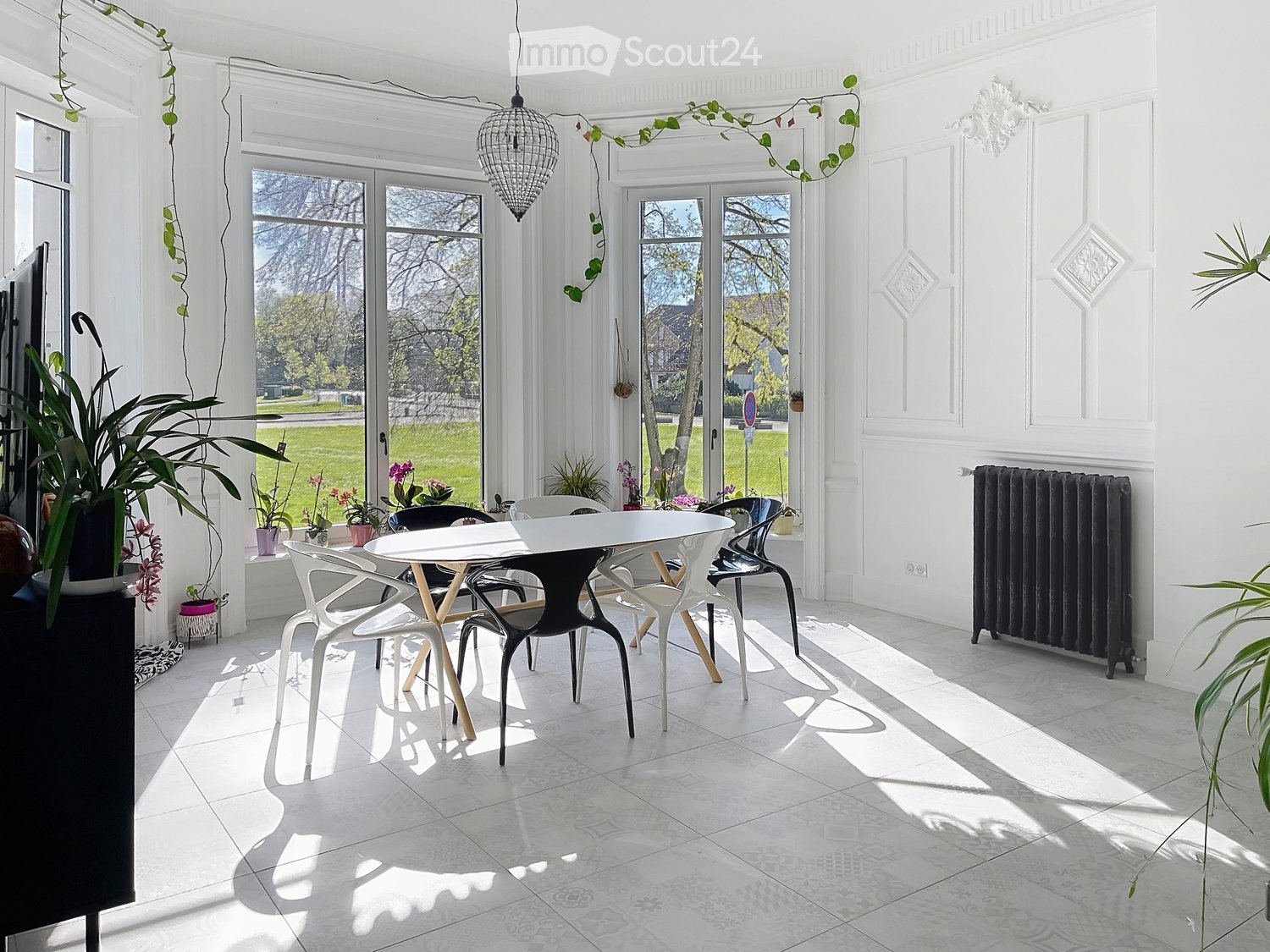 Bright, airy room with large windows, white walls, and a modern dining setup featuring a white table and colorful chairs. Potted plants and a radiator are visible.