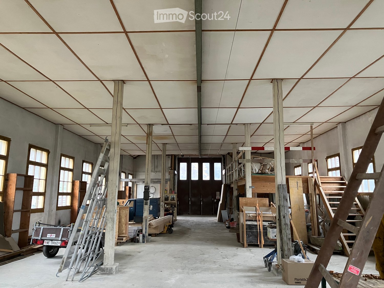 Large, open, unfinished space with exposed wooden beams and metal supports, scattered construction materials, ladders, and storage items.