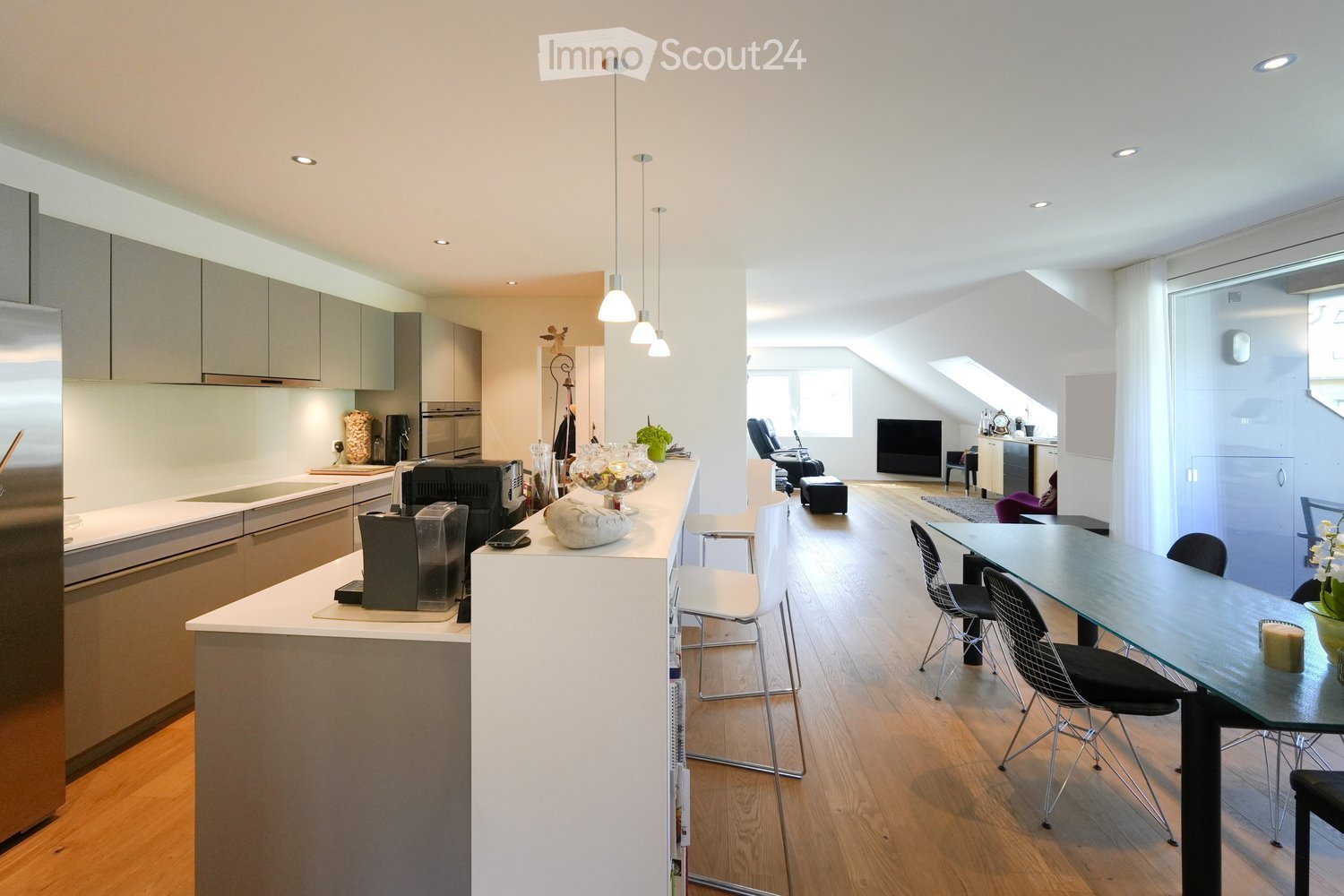 Modern kitchen and living area with white cabinets, stainless steel appliances, a central island, and a dining area with a glass table and chairs. The living area features a TV, a sofa, and a small seating area.