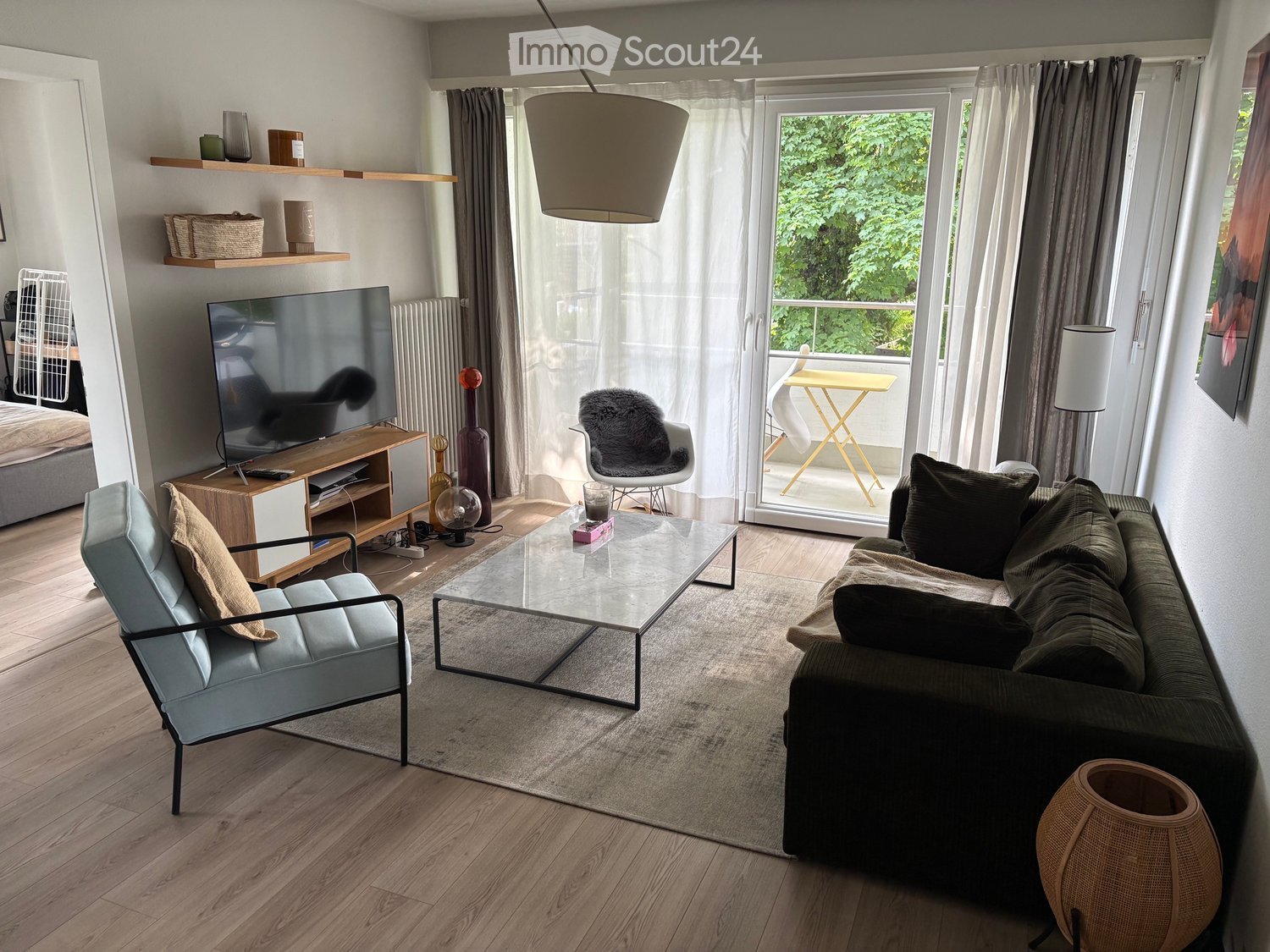 Modern living room with a grey sofa, glass coffee table, light blue armchair, TV stand, pendant lamp, and shelves with decorative items. Large windows with sheer curtains lead to a balcony.