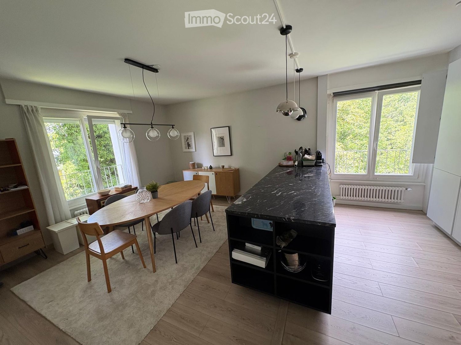 Open-plan dining and kitchen area with wooden dining table, chairs, kitchen island, pendant lights, and large windows overlooking a balcony.