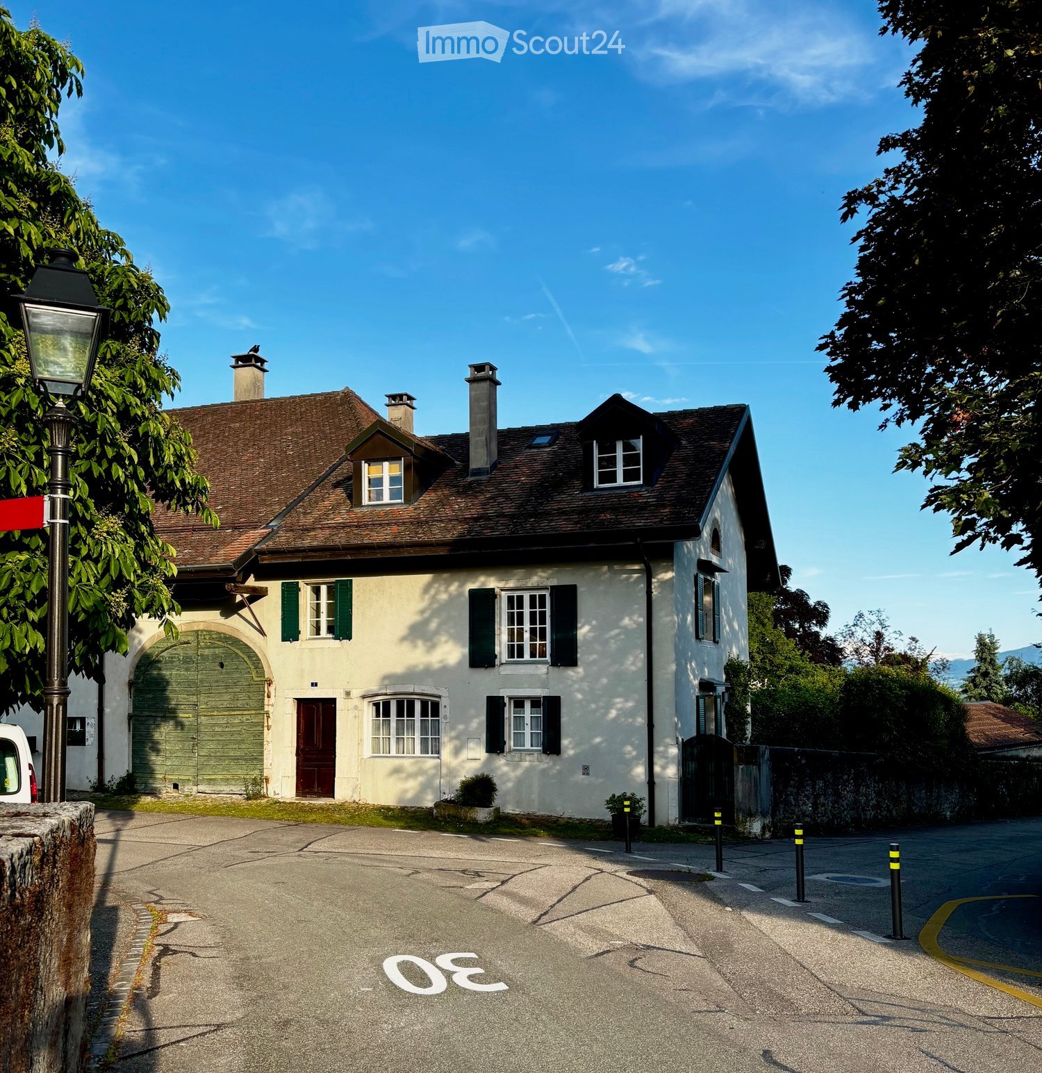 Two-story house with a brown tiled roof, white walls, green shutters, and a green garage door. The house has multiple windows and chimneys, surrounded by trees and a paved driveway.