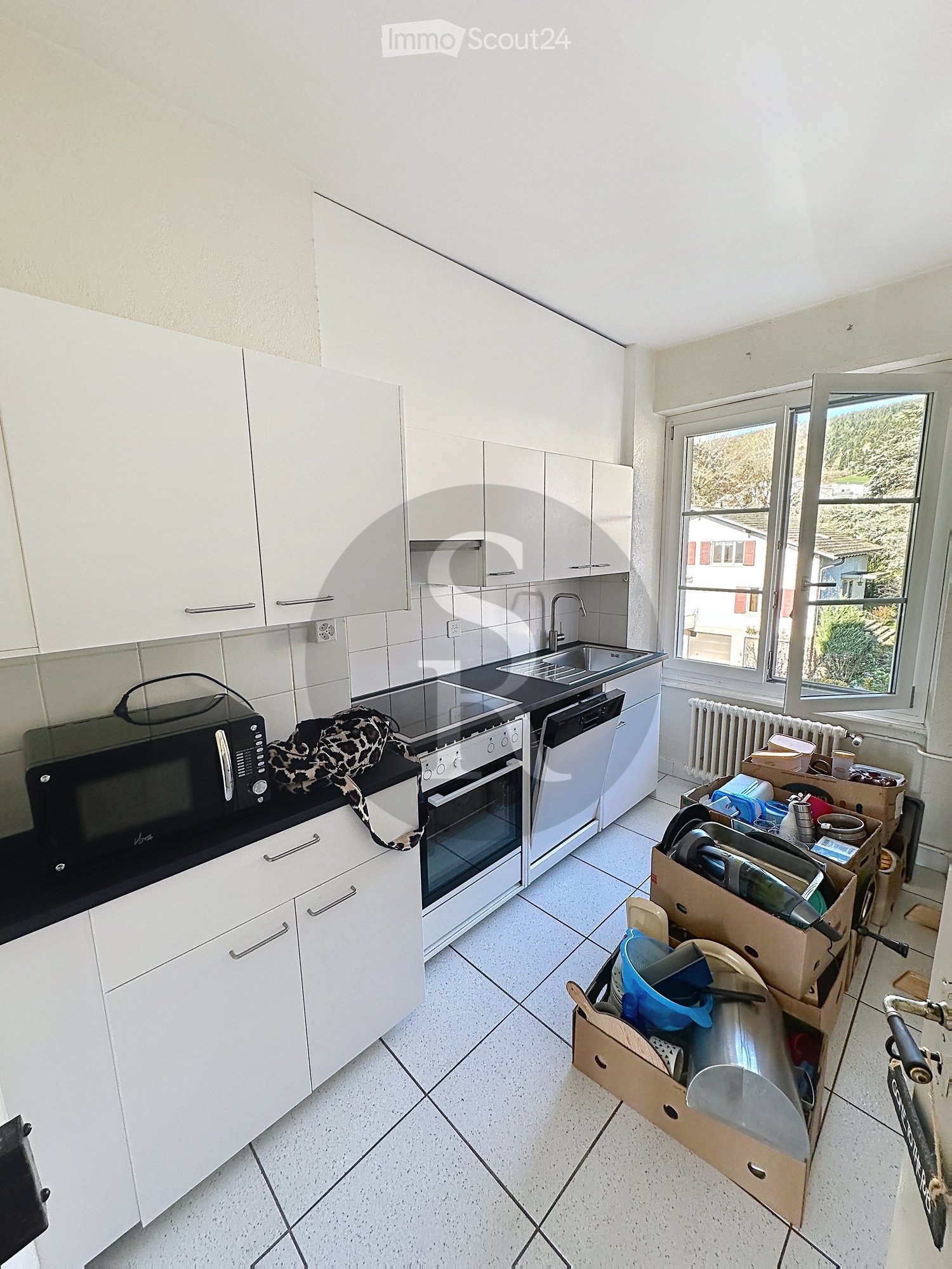 Modern kitchen with white cabinets, a microwave, oven, dishwasher, and sink. There are cardboard boxes with dishes and kitchen items on the floor, and a bicycle is partially visible on the right side.