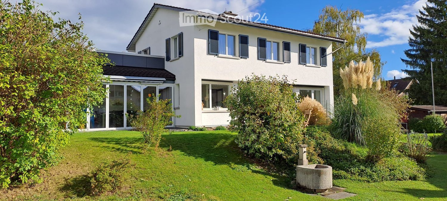 Two-story white house with multiple windows, shutters, and a modern glass-walled extension. The house is surrounded by a well-maintained garden with various plants and a stone well.