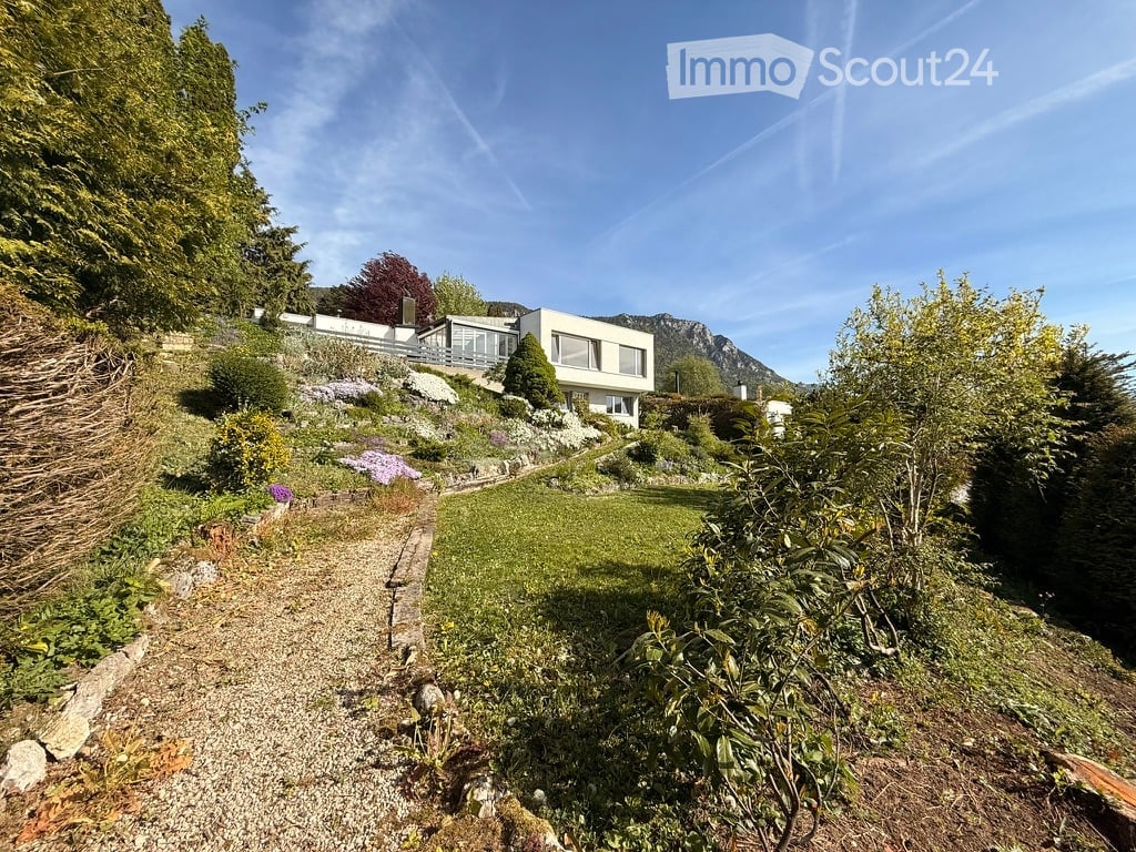 A modern house with a raised ground floor, surrounded by a landscaped garden with a gravel path, lush greenery, and a scenic mountain view in the background.