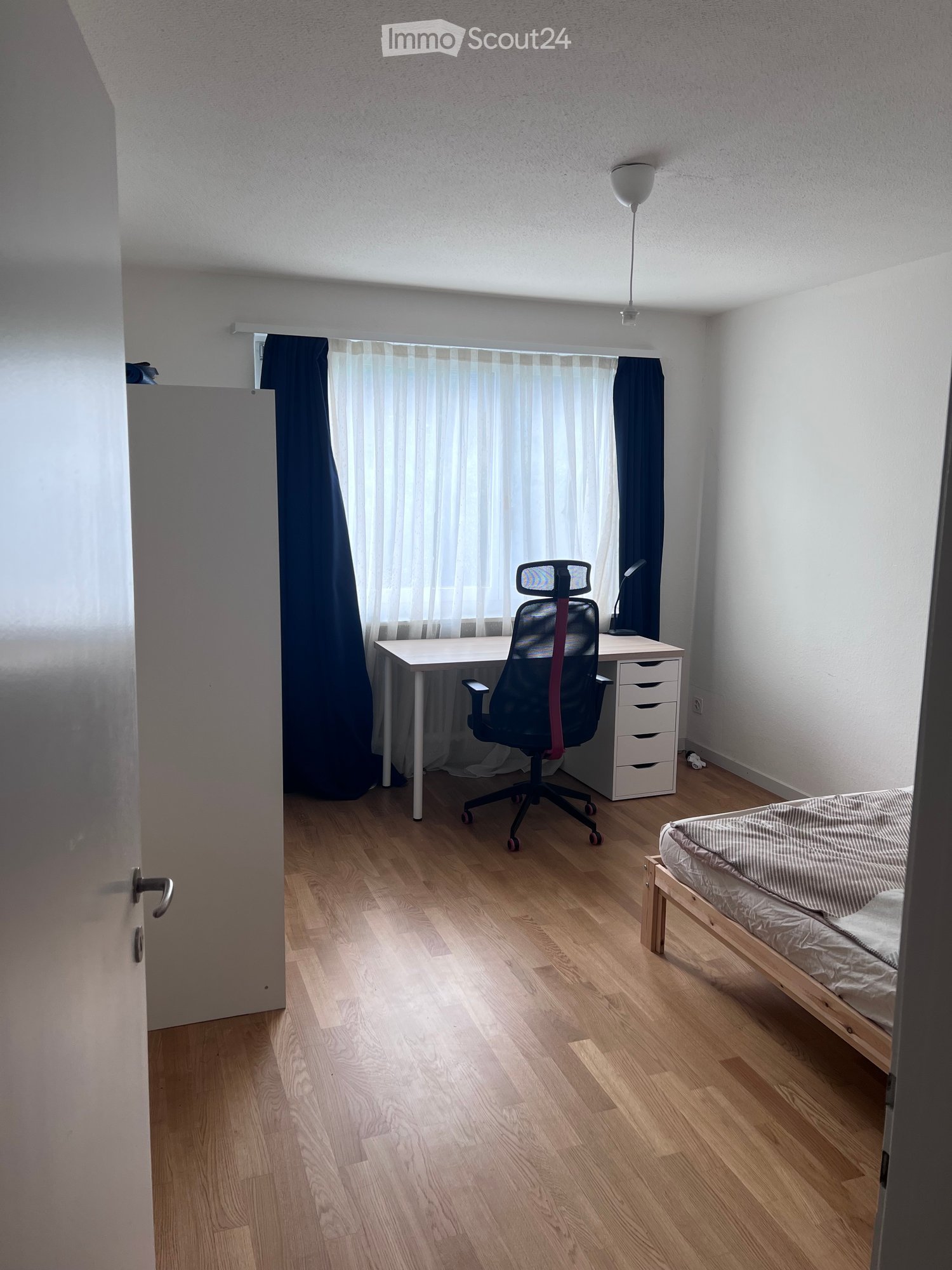 A modern, minimalist bedroom with a wooden floor, a bed with a grey duvet, a desk with a chair, and a window with curtains.