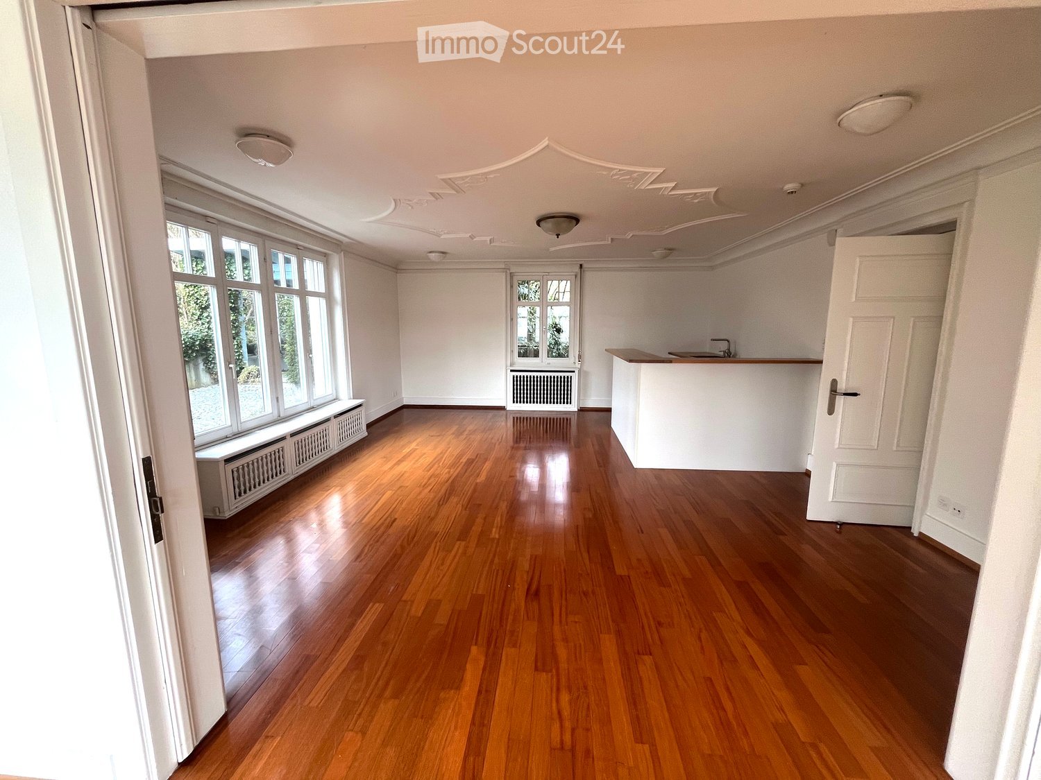 Empty room with wooden flooring, white walls, large windows, and a small counter area with a sink.