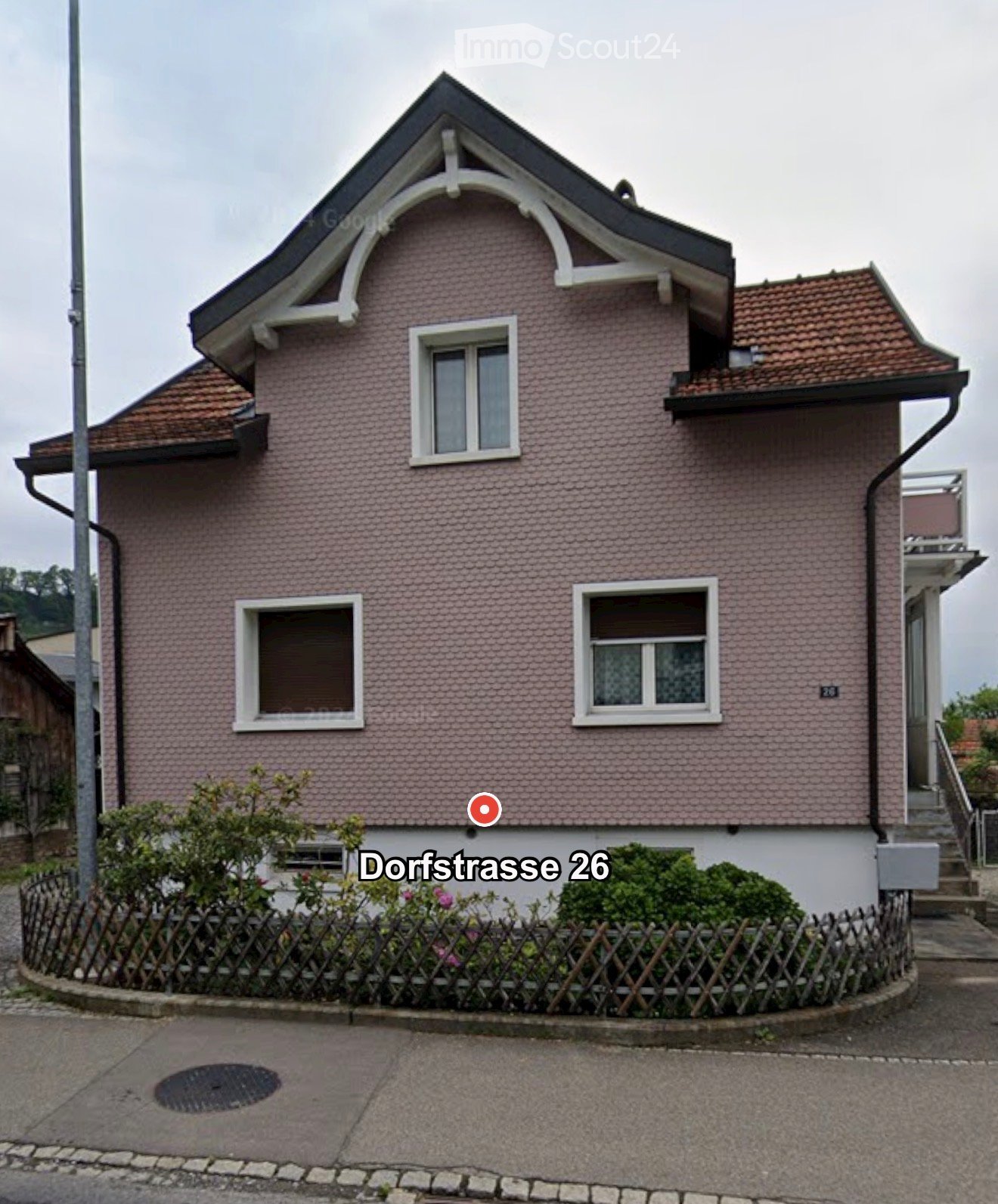 Two-story house with pink tiled exterior, white-framed windows, and a decorative roofline. The house features a small balcony on the right side and is surrounded by a wooden fence with greenery.
