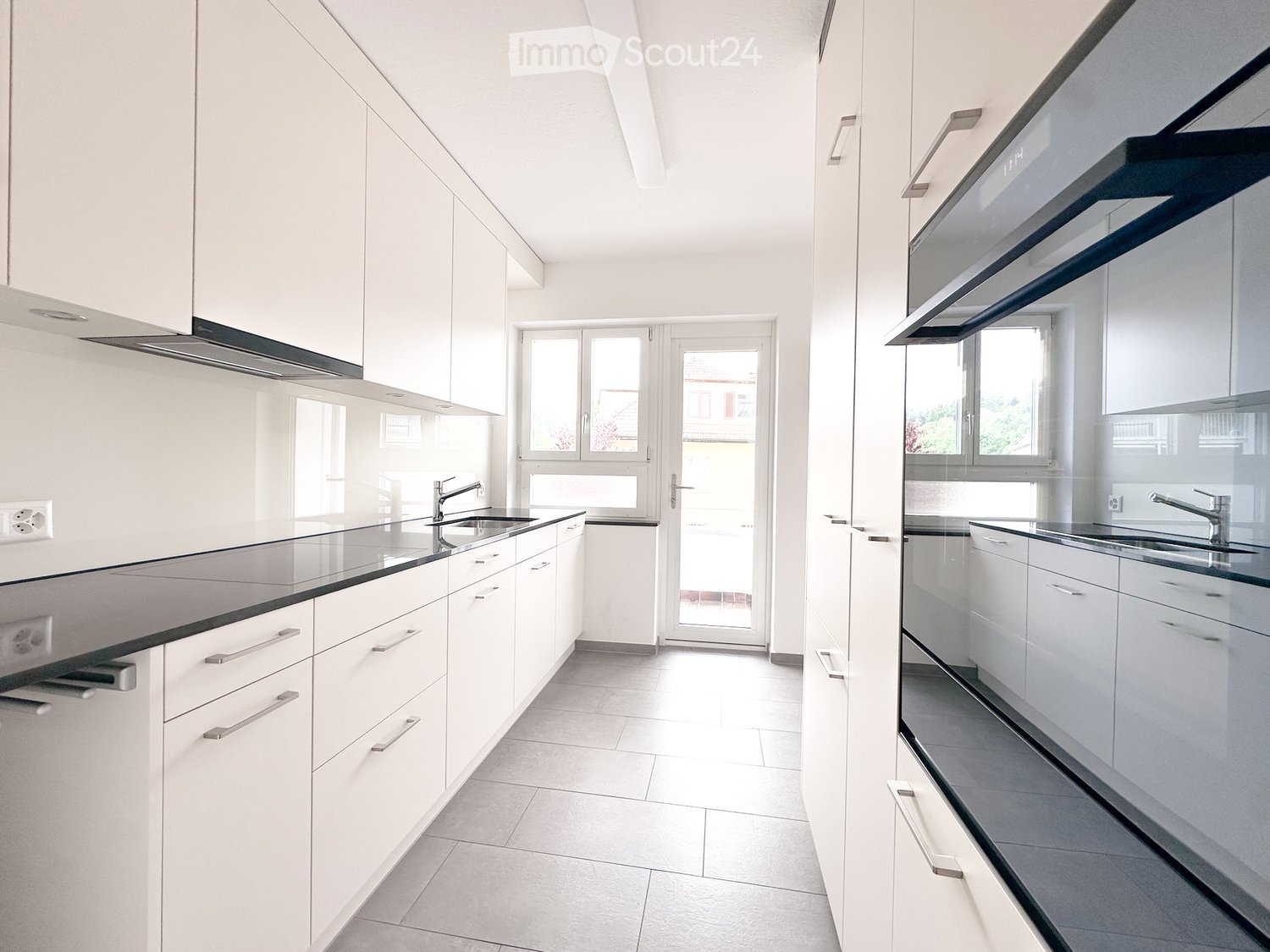 Modern, minimalist kitchen with white cabinets, sleek countertops, and built-in appliances. Features a large window and a glass door leading to an outdoor area.