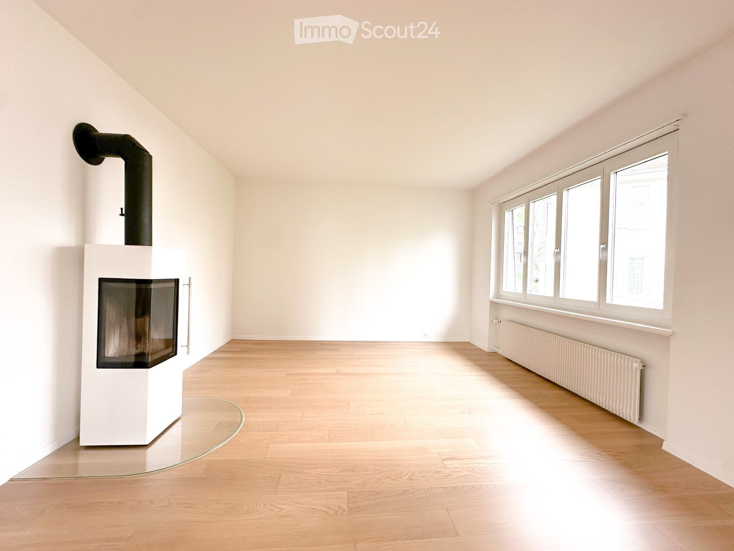 Empty room with light wooden flooring, white walls, large windows, and a modern white fireplace with a black chimney.