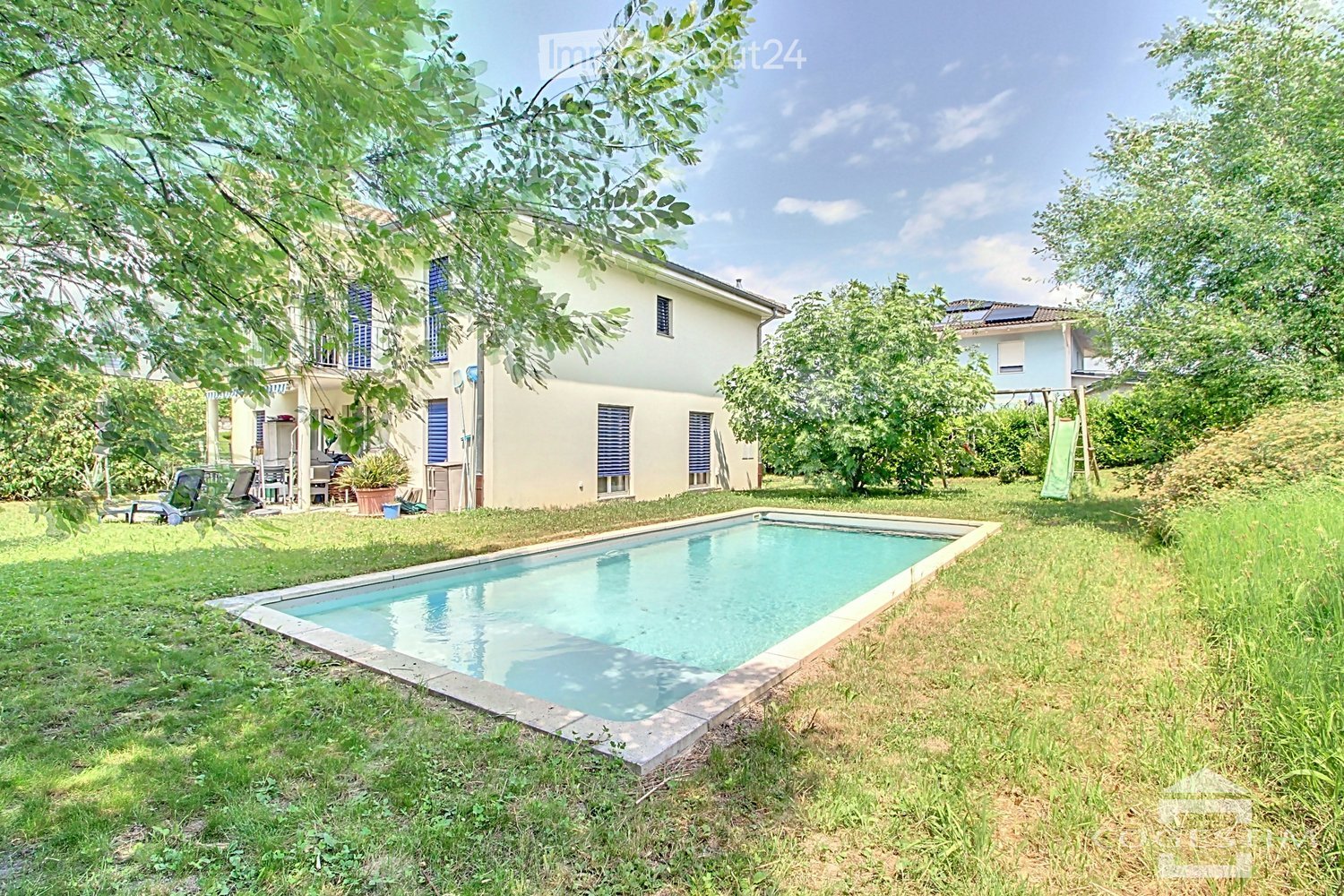 A sunny garden area with a rectangular swimming pool, surrounded by green grass and trees. A two-story house with blue shutters and a balcony is visible in the background, along with outdoor furniture and a swing set.