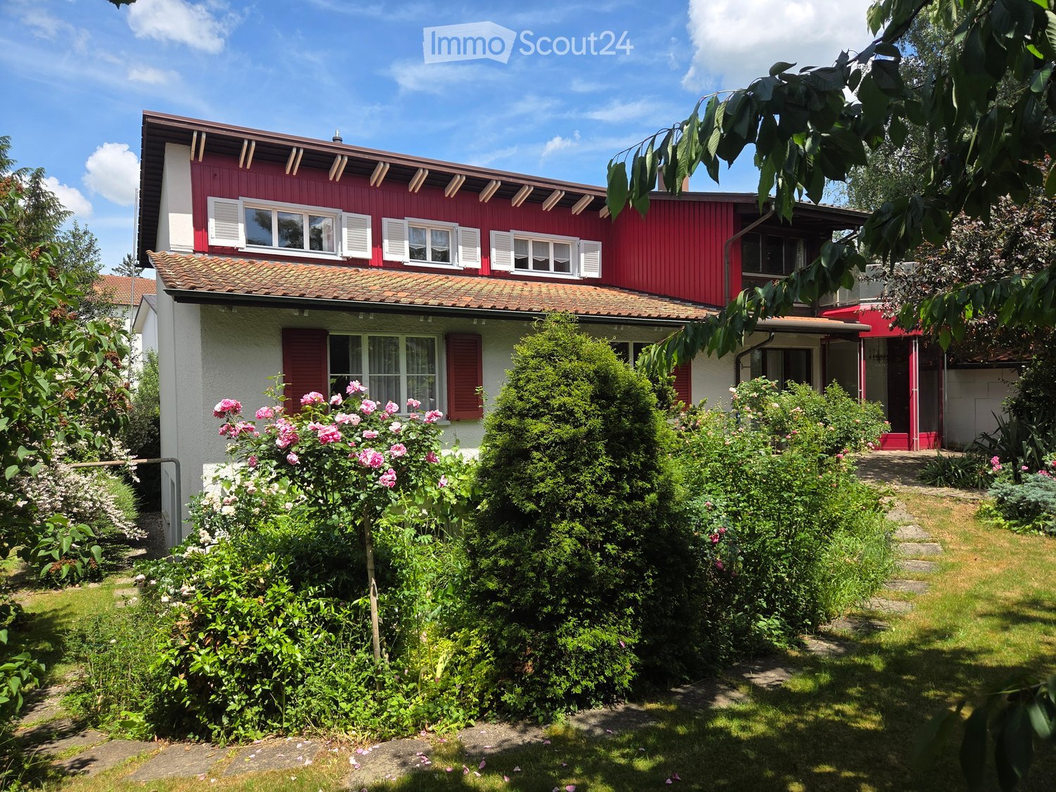 Two-story red and white house with a tiled roof, multiple windows, and a well-maintained garden with various plants and flowers.