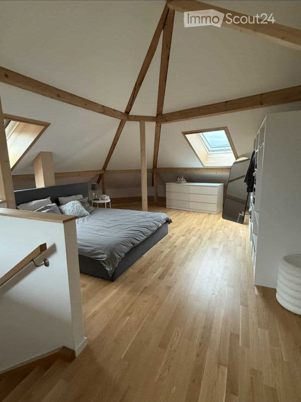 Bright, modern attic bedroom with wooden floors, slanted wooden ceiling beams, skylight windows, and contemporary furnishings including a bed, nightstand, and dresser.
