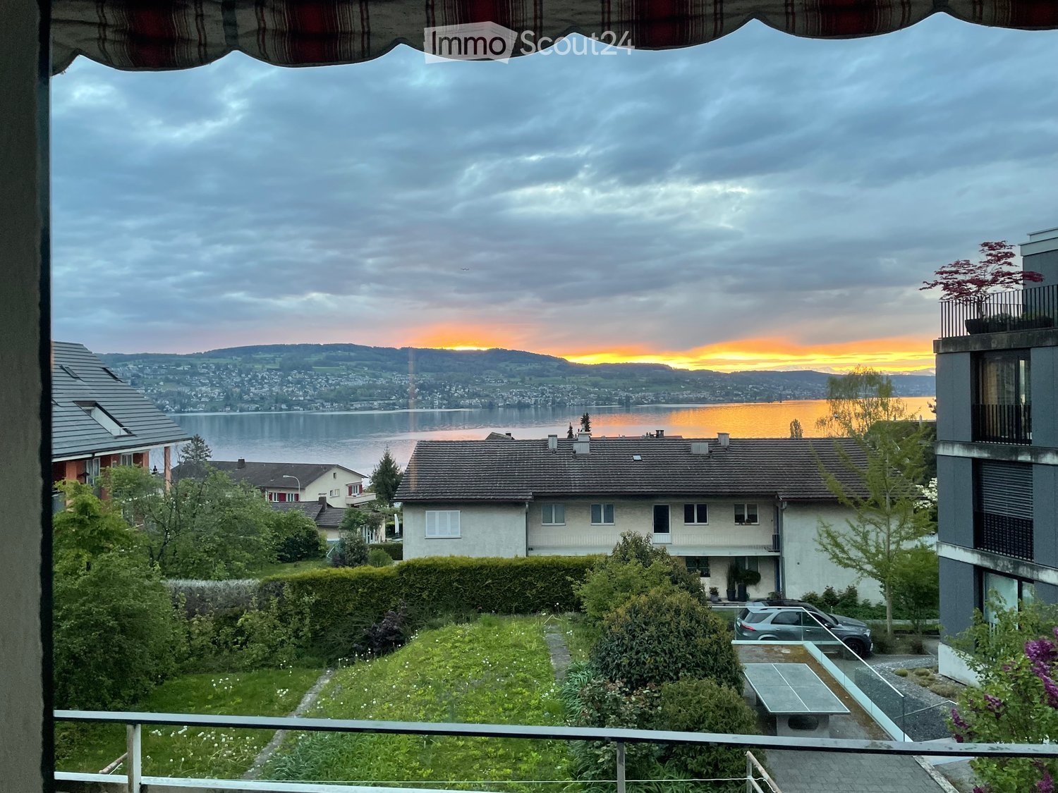 View from a balcony with a bench, overlooking a car parked in front of a house, a lake, and a sunset.