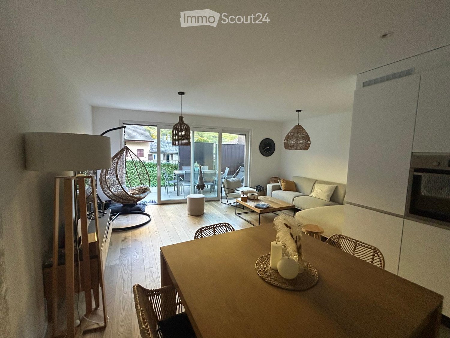 Open-plan living and dining area with wooden flooring, large sliding glass doors leading to a patio, a sofa, coffee table, wicker hanging chair, dining table with chairs, and decorative items.