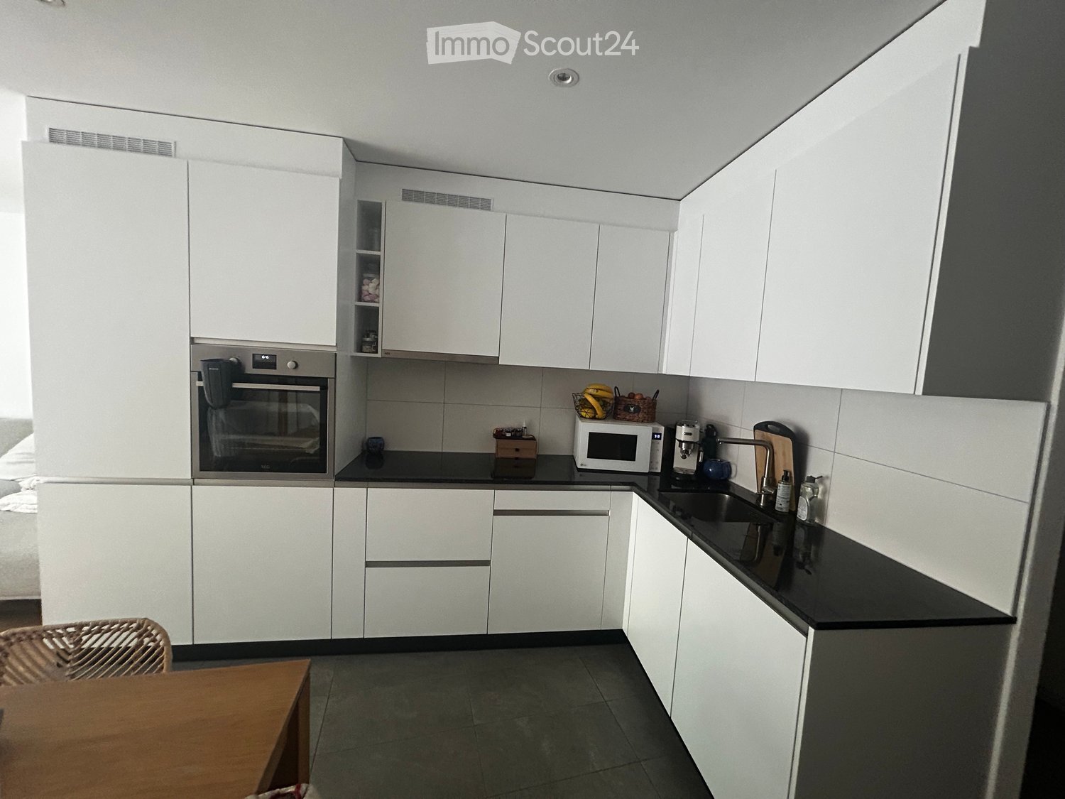 Modern kitchen with white cabinets, black countertops, and stainless steel appliances including an oven and microwave. A small table and chair are visible in the foreground.