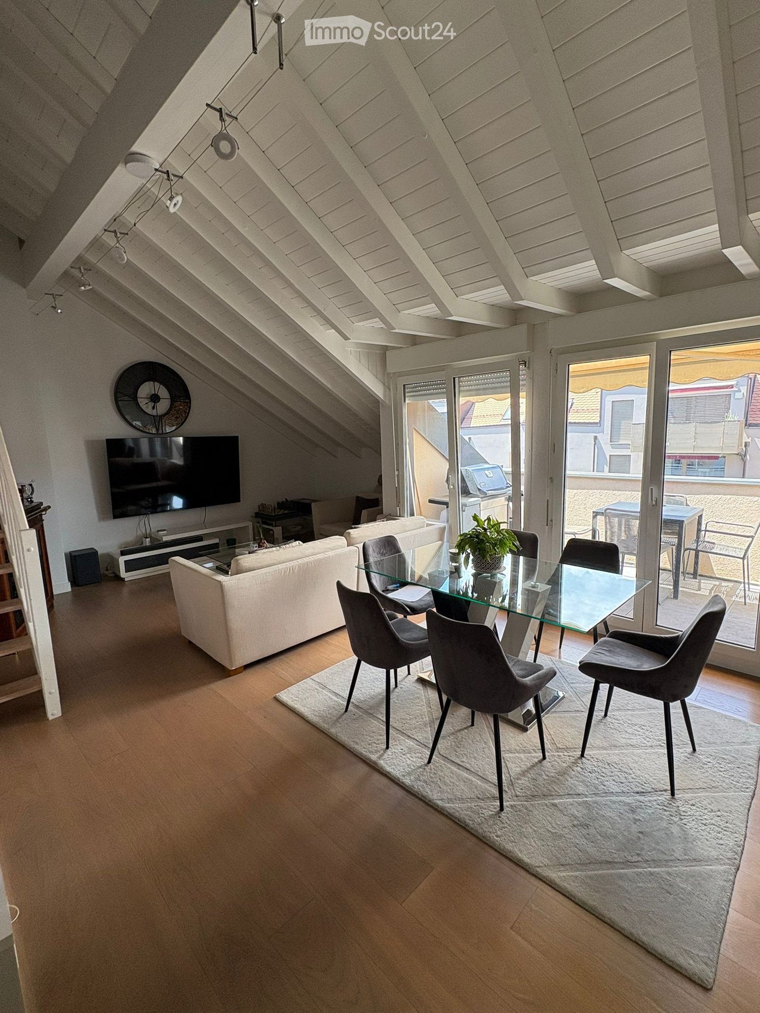 Modern living and dining area with wooden flooring, white walls, and a slanted ceiling. Features a white sofa, glass dining table with four chairs, a television, and a clock on the wall. A large window with a view of the balcony and outdoor area.