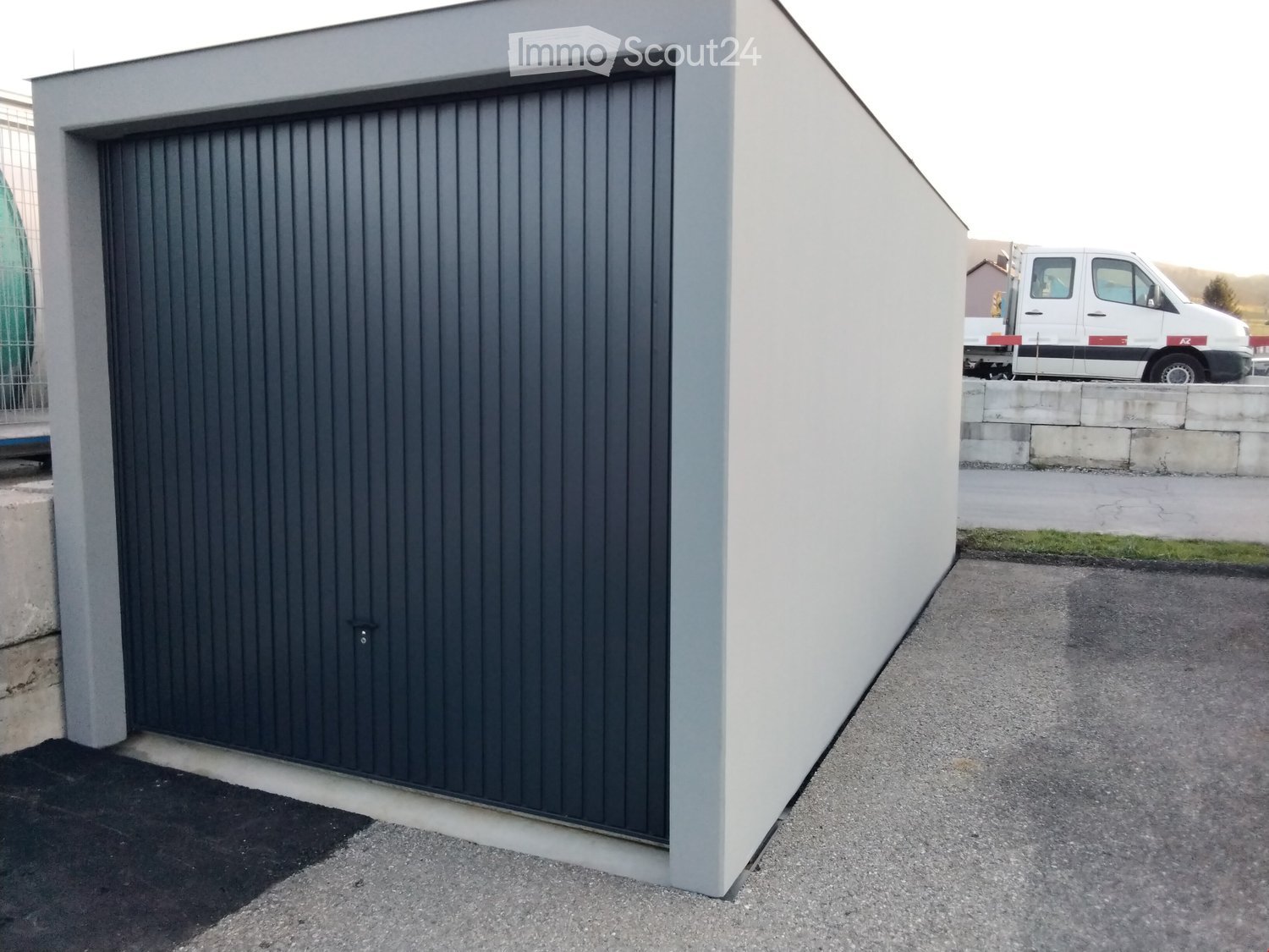 Modern building entrance with a large, dark grey garage door and a white wall. A white van is parked in the background.