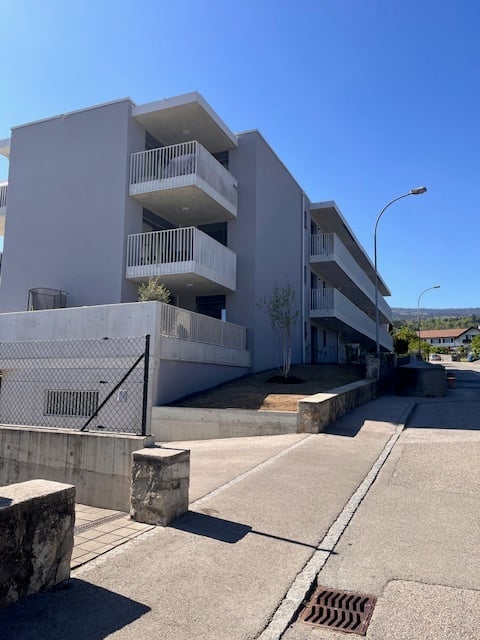 Modern multi-story apartment building with white balconies and clean lines, situated on a raised ground floor with a paved walkway.