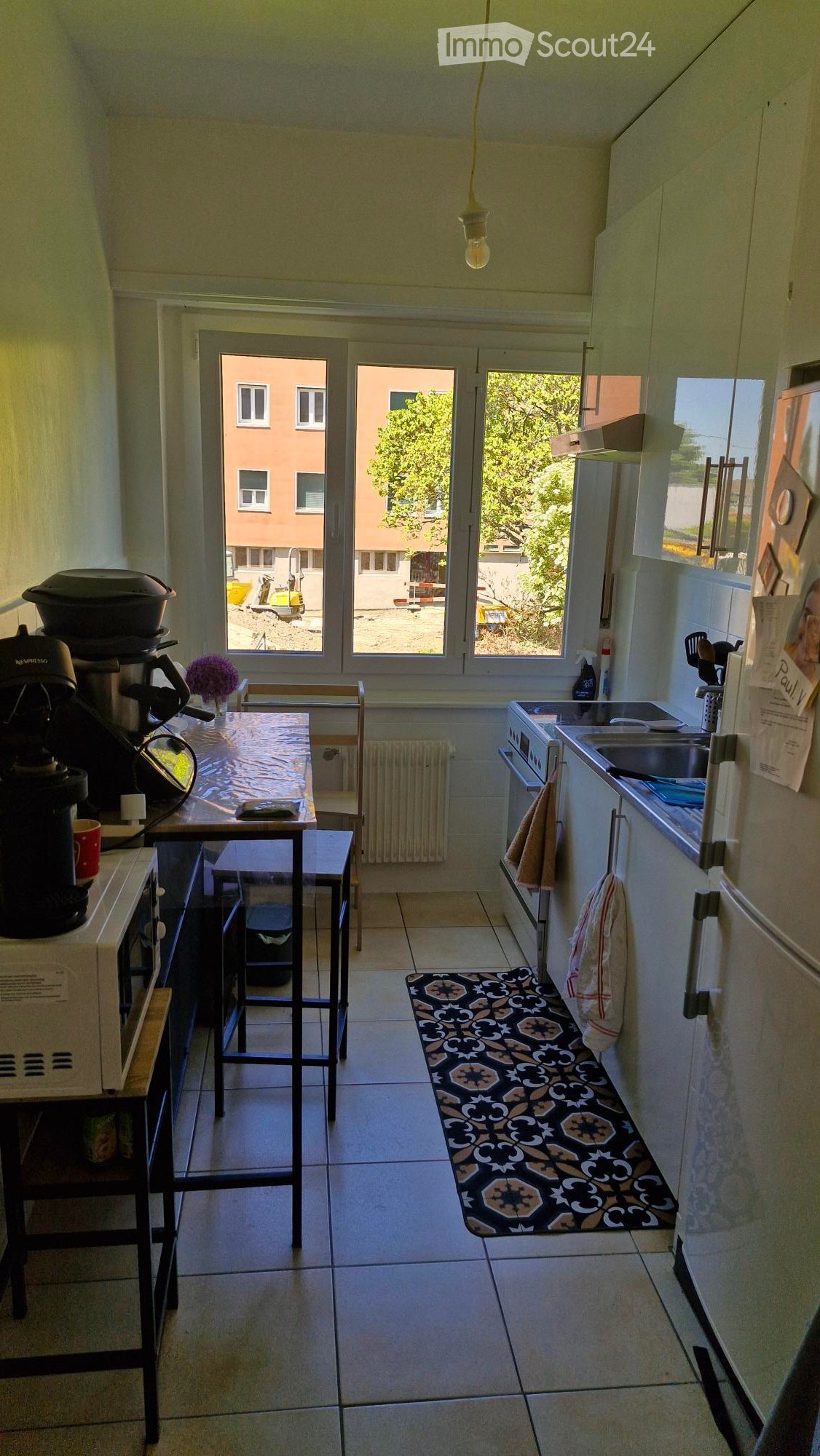 The kitchen features a coffee maker, a small table with a stool, an oven, a sink, and a refrigerator. There is a patterned rug on the floor and a flower vase on the table.