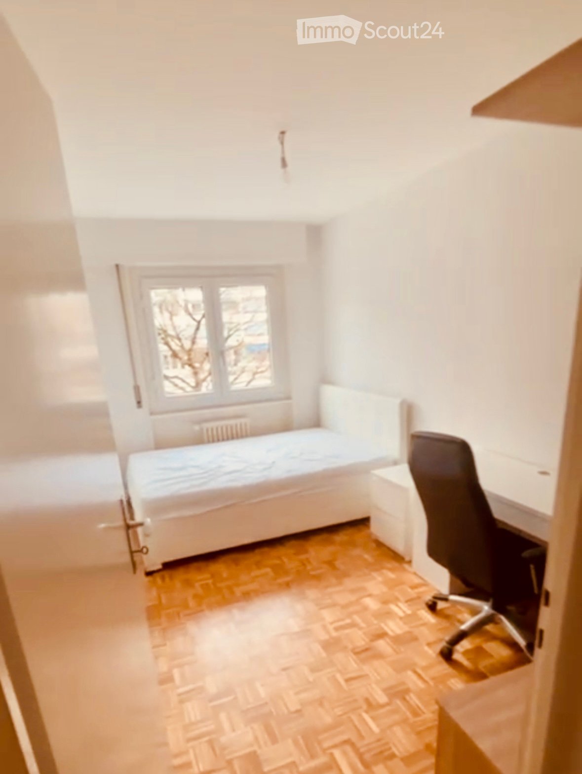 A small, furnished room with a single bed, a desk, and a chair. The room has wooden flooring and a window with a view of trees.