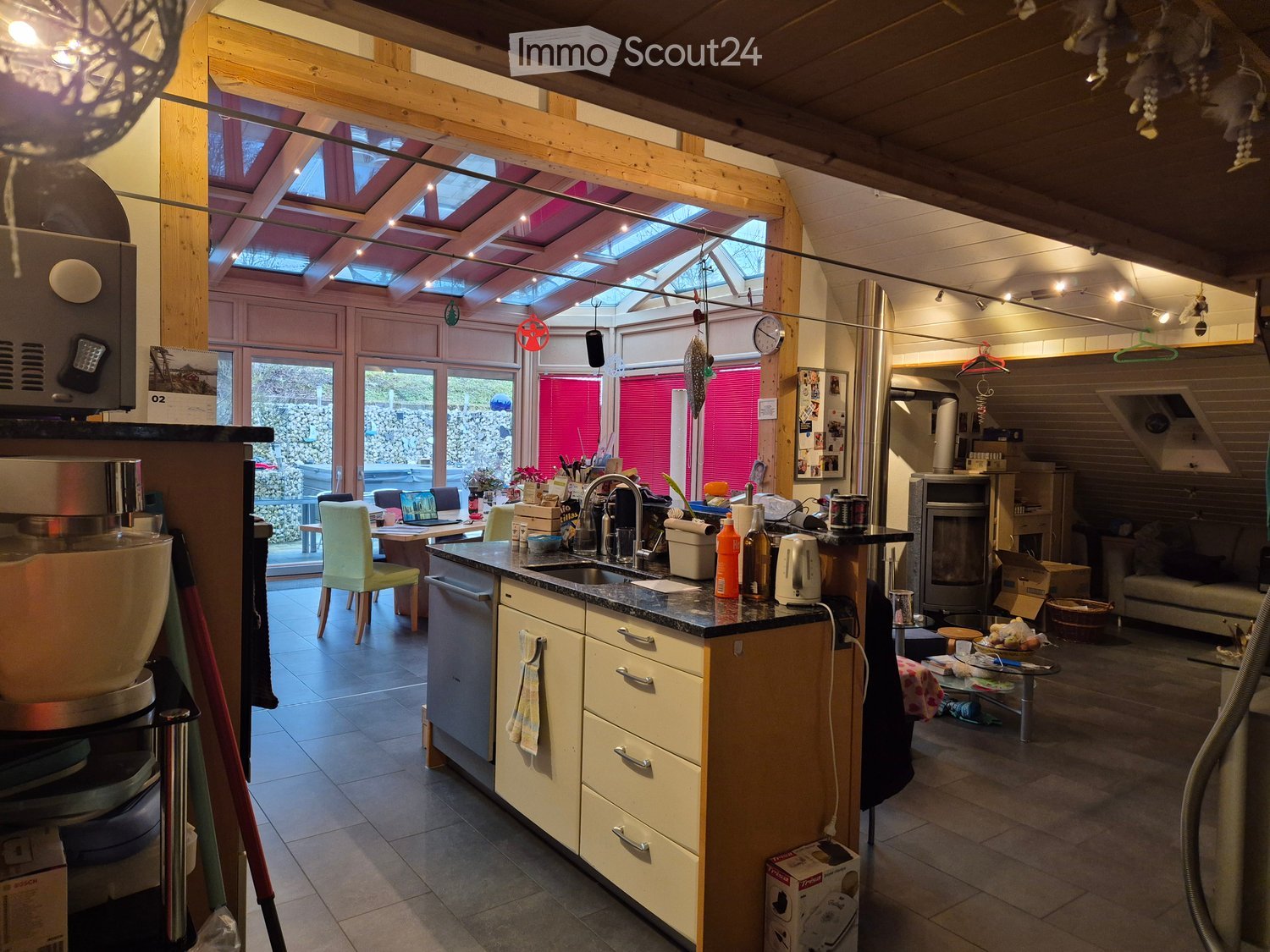 Open-plan kitchen and living area with wooden beams, skylights, and modern appliances. Includes a coffee maker, sink, dishwasher, refrigerator, oven, and a living area with a sofa and fireplace.