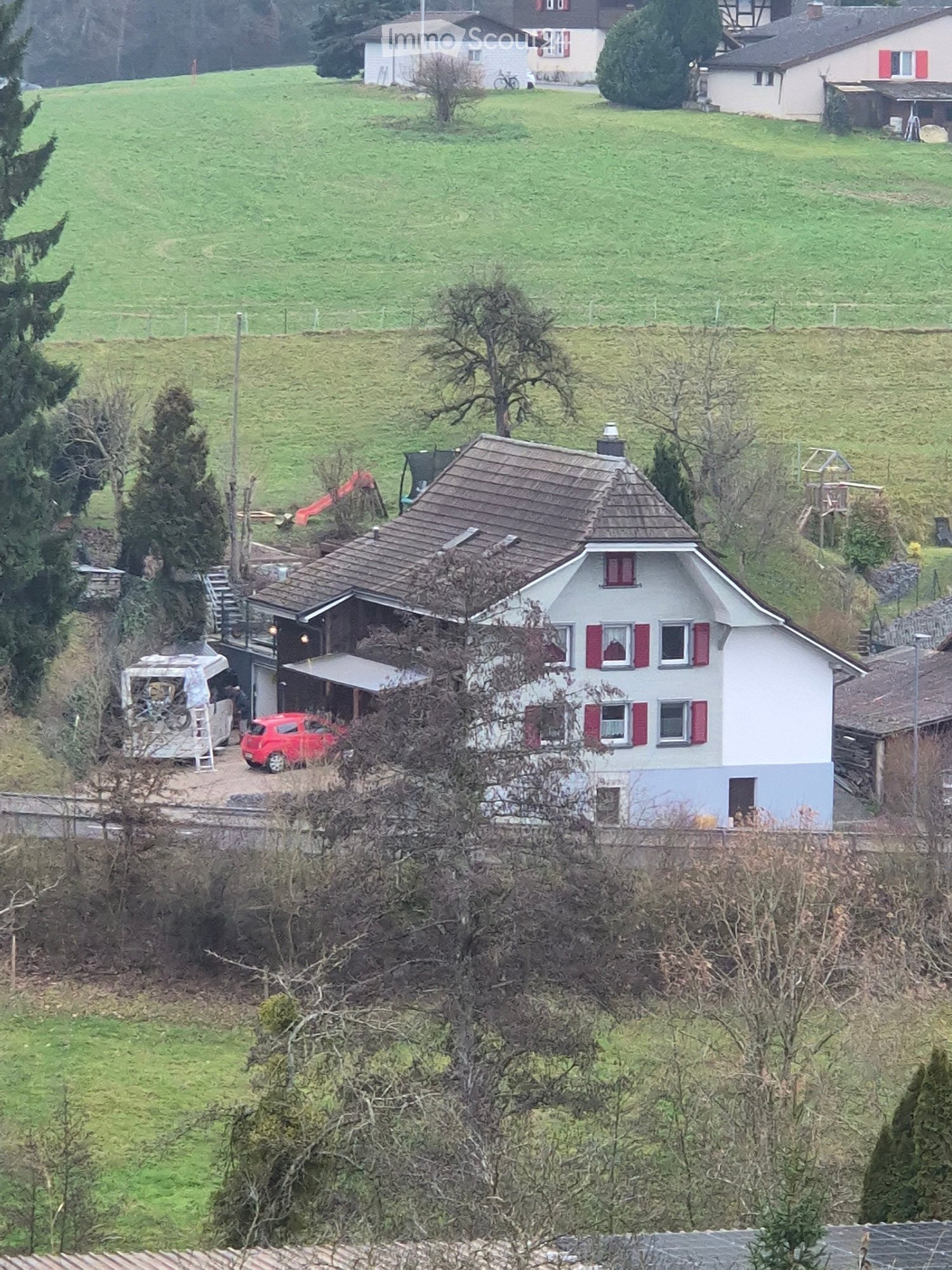 A two-story house with a brown tiled roof and red-framed windows, situated on a hillside with surrounding greenery and neighboring houses visible in the background.