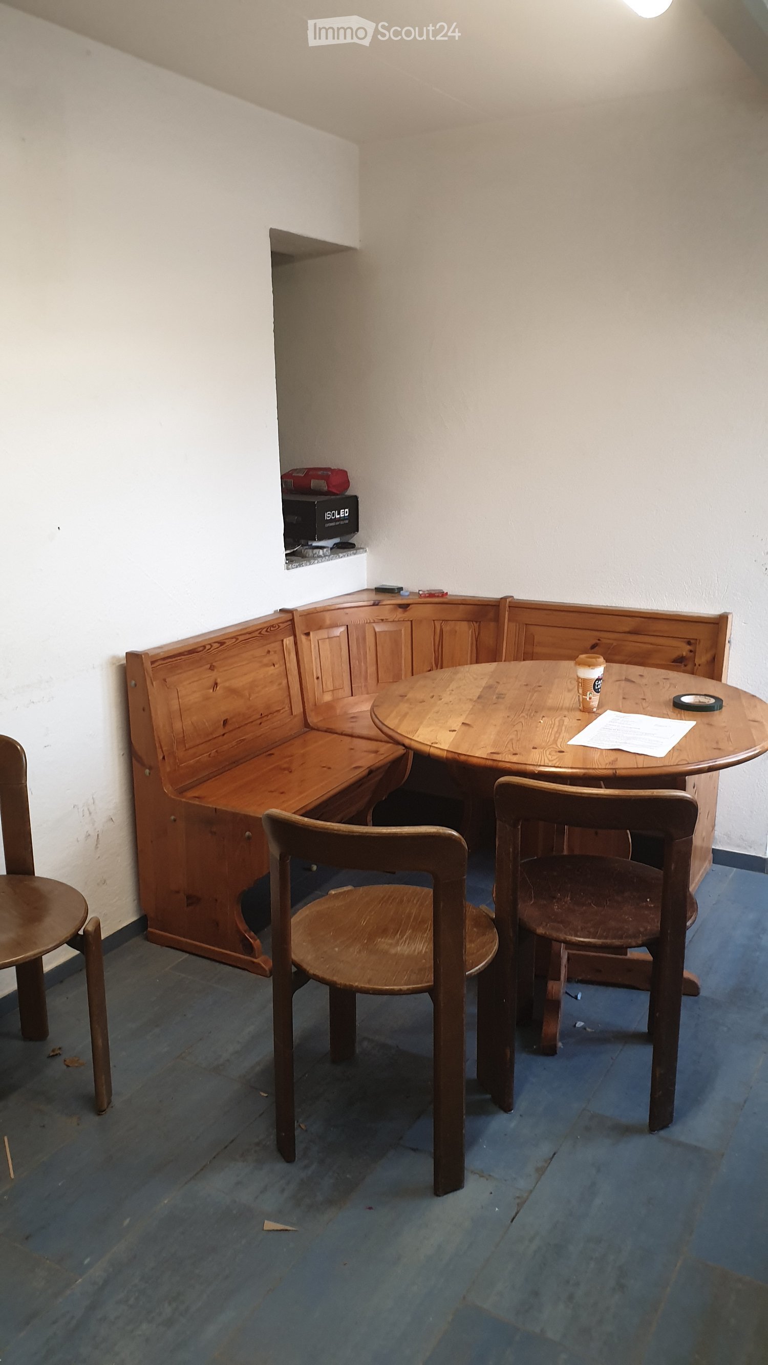 A small, cozy room with a round wooden table, four wooden chairs, and a built-in wooden bench along one wall. The room has white walls and a blue wooden floor.