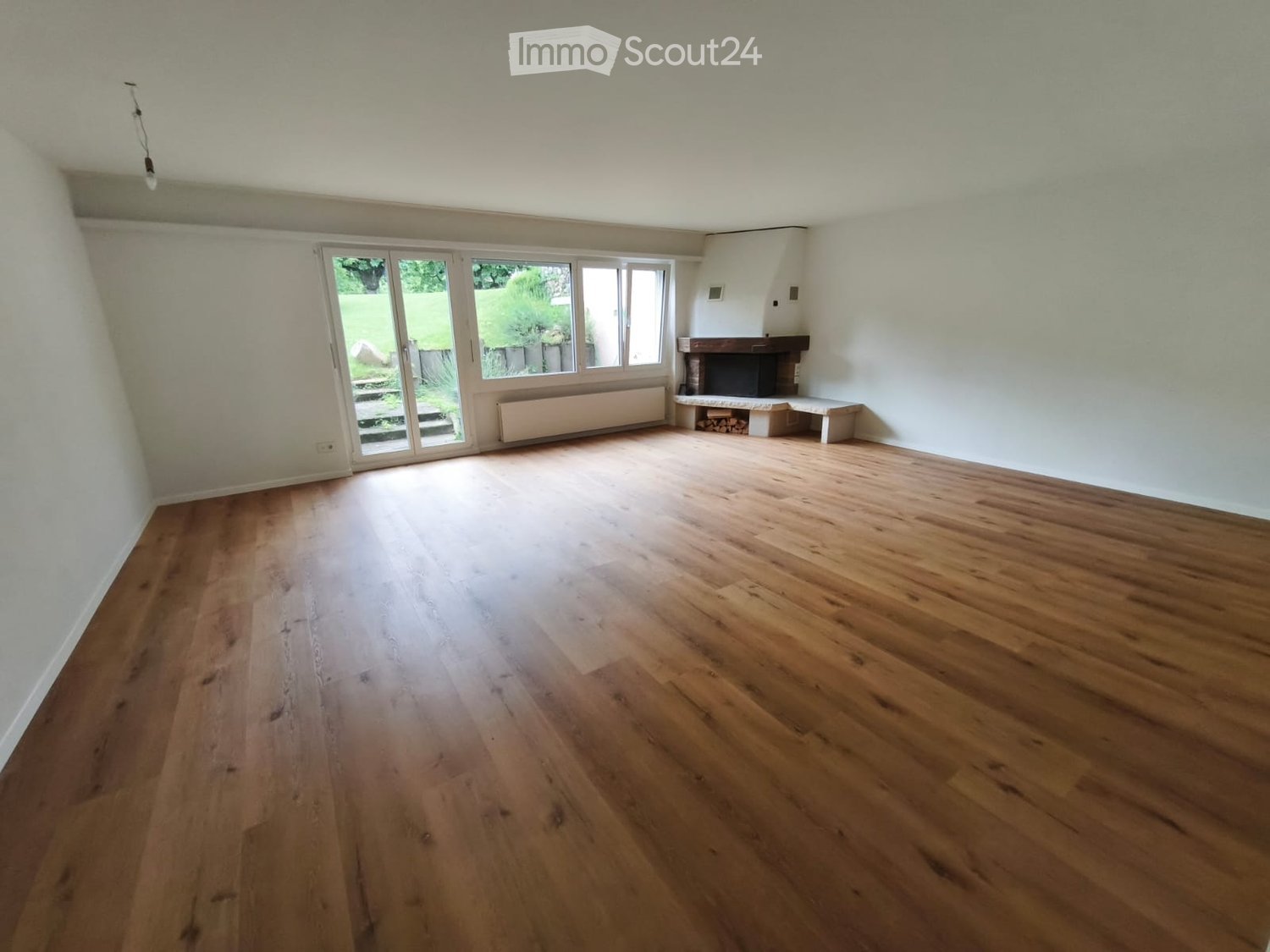 Empty room with wooden flooring, white walls, large sliding glass doors leading to a garden, and a fireplace with a bench.
