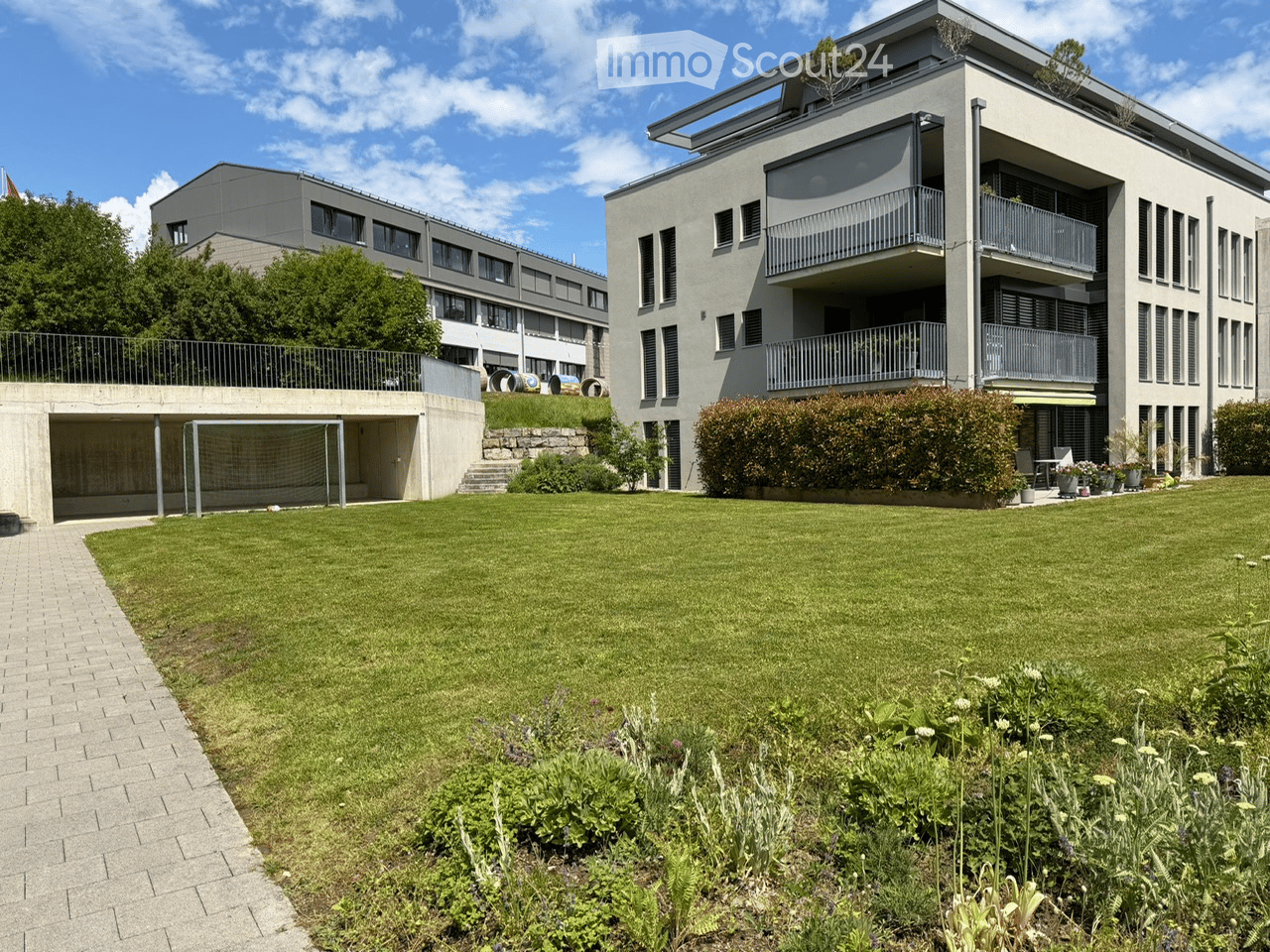 Modern multi-story building with balconies, surrounded by a well-maintained lawn and garden area. Includes a covered garage and raised ground floor.