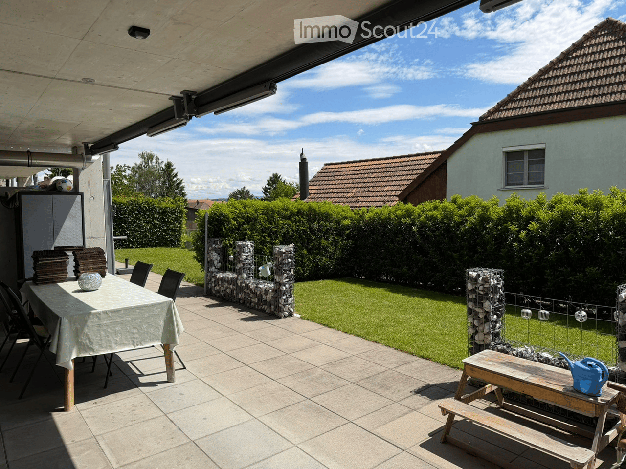 Outdoor patio with a dining table, chairs, a wooden bench, and a watering can, surrounded by a well-maintained lawn and hedges.