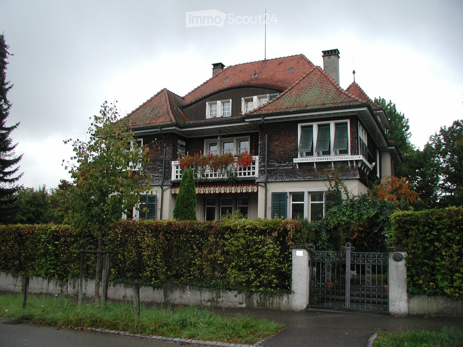 Two-story house with red-tiled roof, multiple windows, balconies, and surrounding greenery.