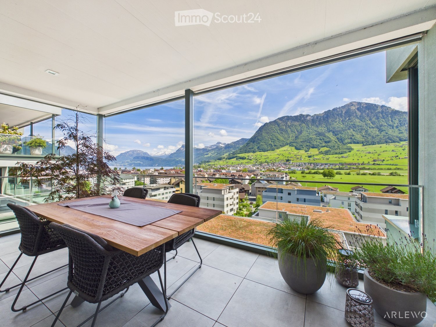 Balcony with a wooden dining table, four wicker chairs, a vase, and potted plants, offering a scenic mountain view.