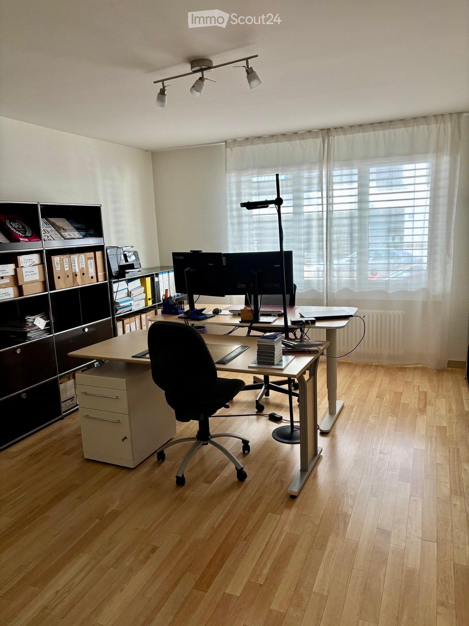 A well-lit office space with a wooden floor, a large desk with a chair, a laptop, a screen, books, a shelf, and a lamp.