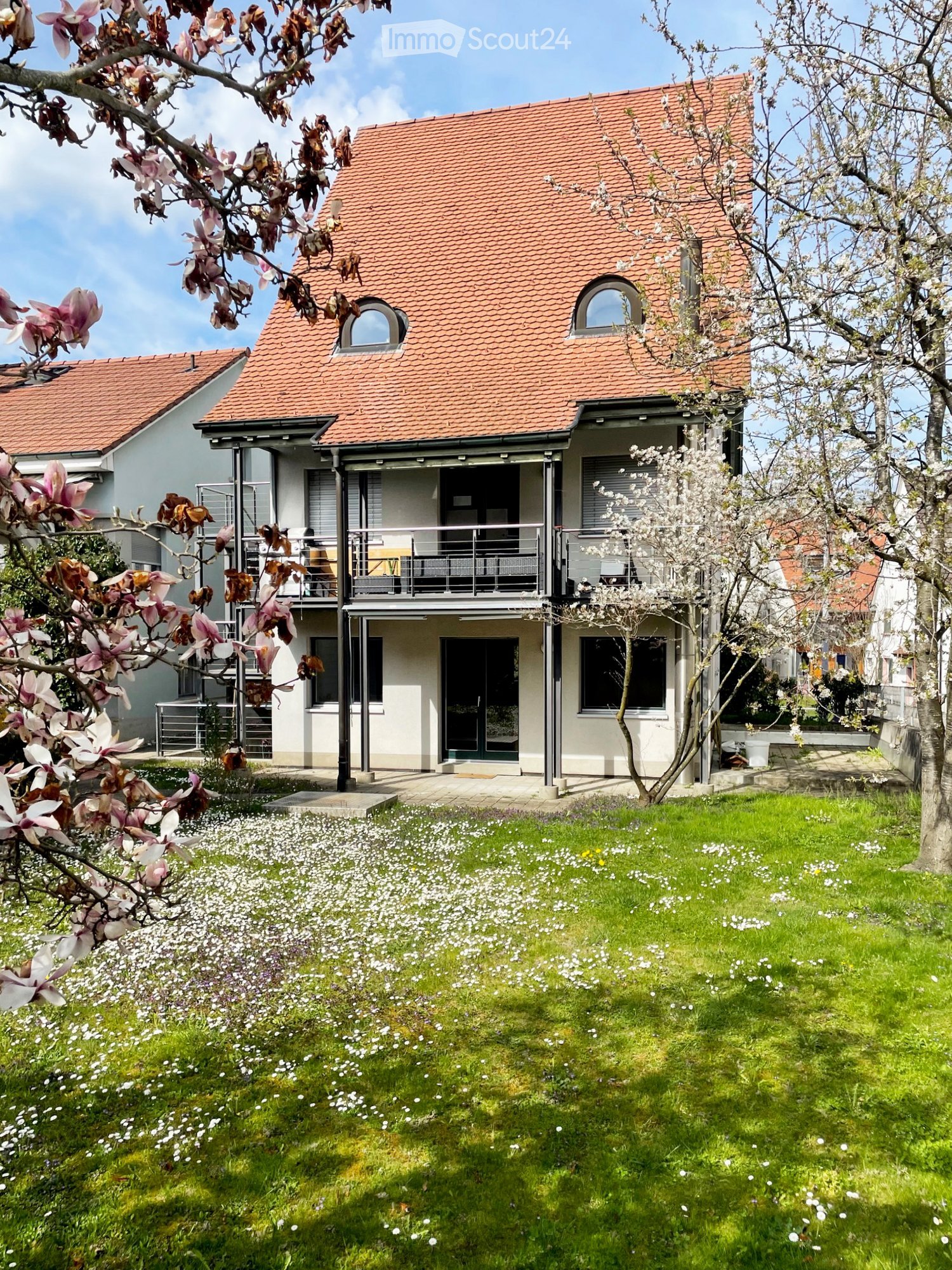 Two-story modern house with red-tiled roof, large windows, and a balcony. The front yard features a well-maintained lawn with white flowers and blooming trees.