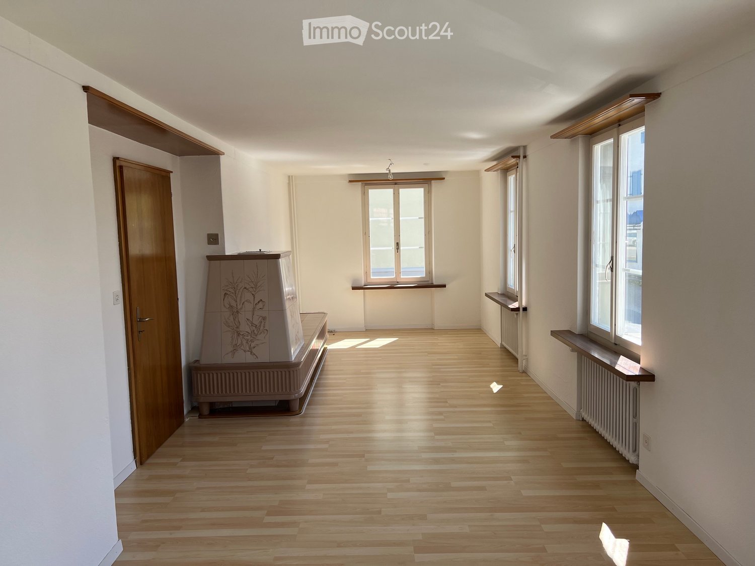 Empty hallway with wooden flooring, white walls, and large windows allowing natural light. Features include a wooden door on the left and built-in window sills on the right.