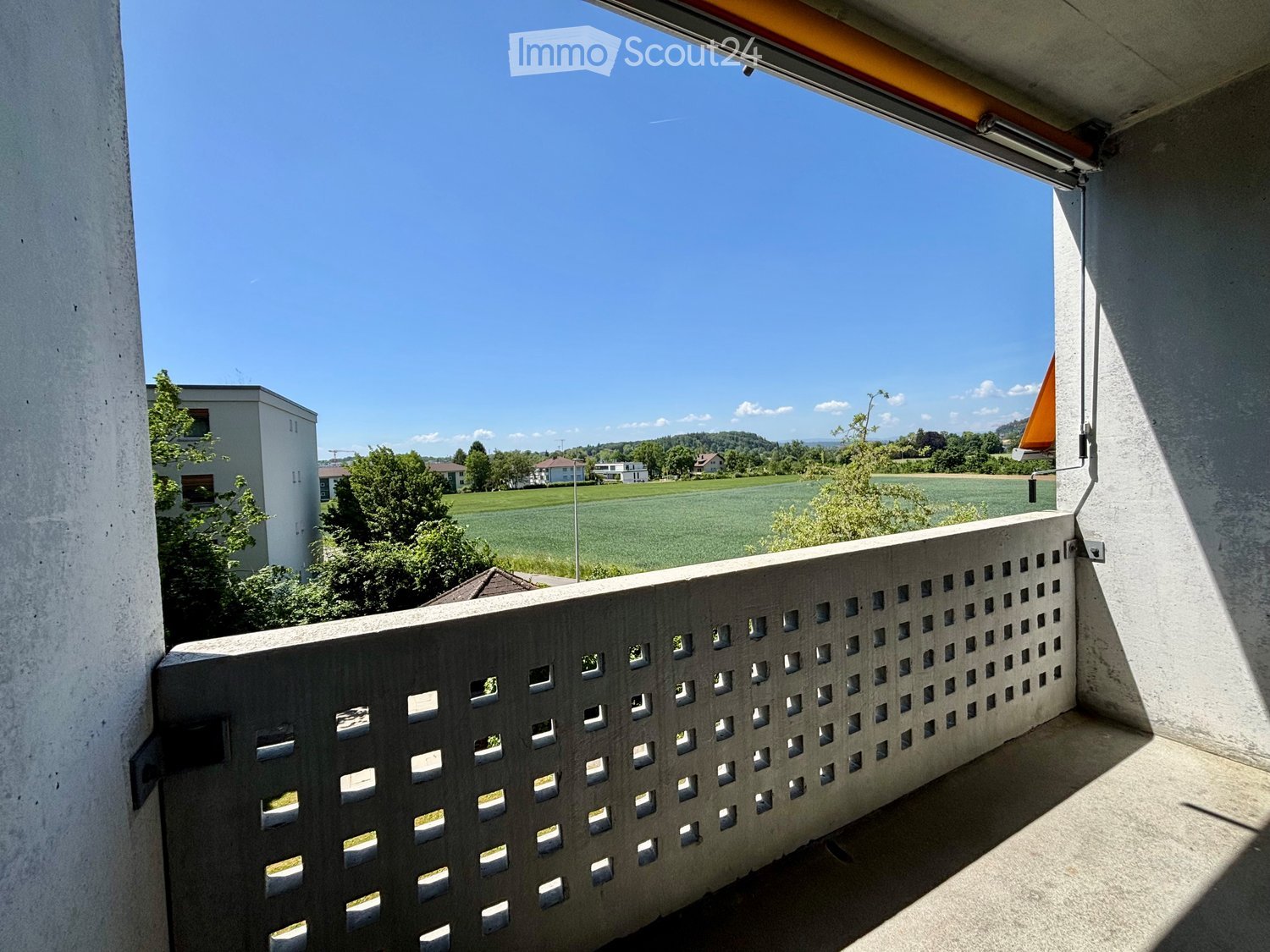 Balcony with a perforated concrete railing, overlooking a green field and distant buildings.