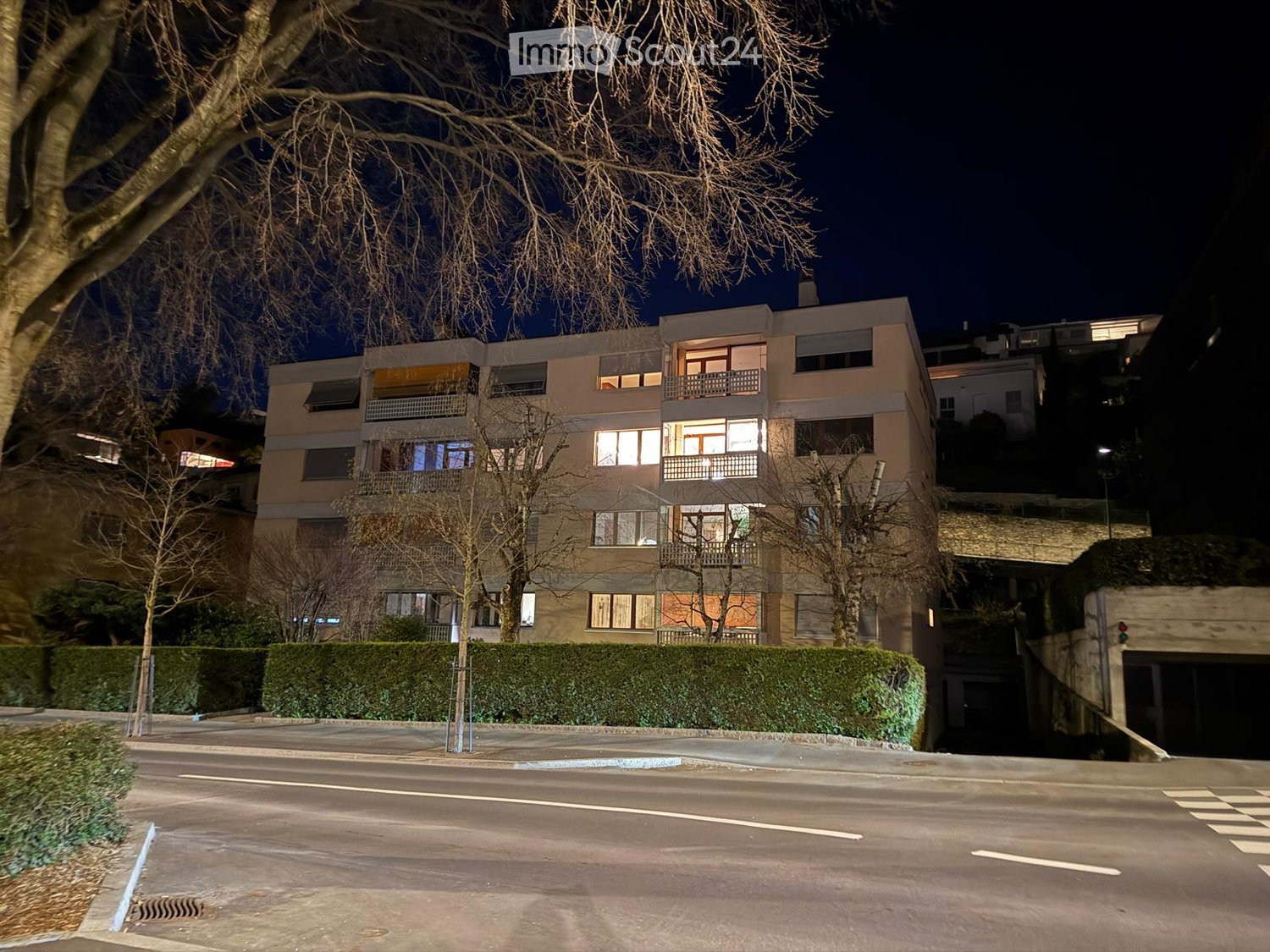 Multi-story residential building with illuminated windows and balconies, surrounded by trees and a hedge, with a street in front.