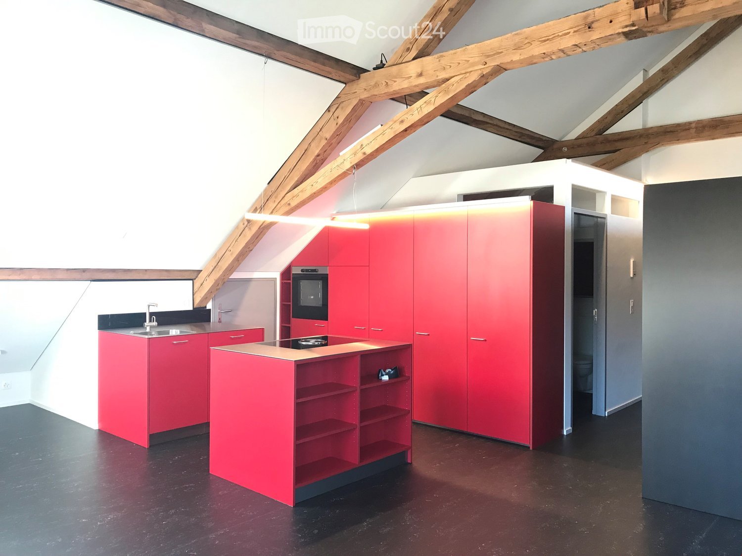 Modern kitchen with red cabinets, stainless steel appliances, and a dark wooden floor. Features a central island with open shelving and a built-in oven.