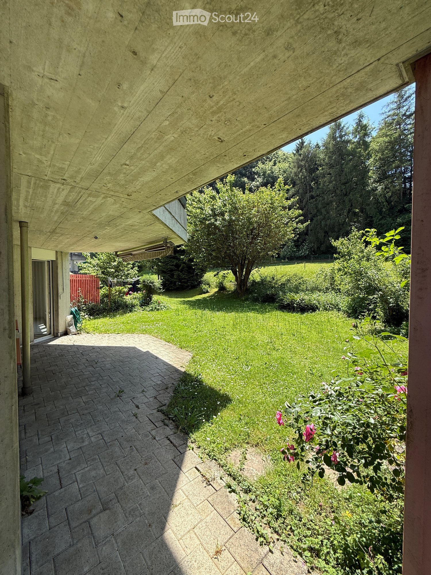 A well-maintained garden with a paved patio, lush green grass, flowering bushes, and trees, surrounded by a modern building with a concrete roof and glass doors.