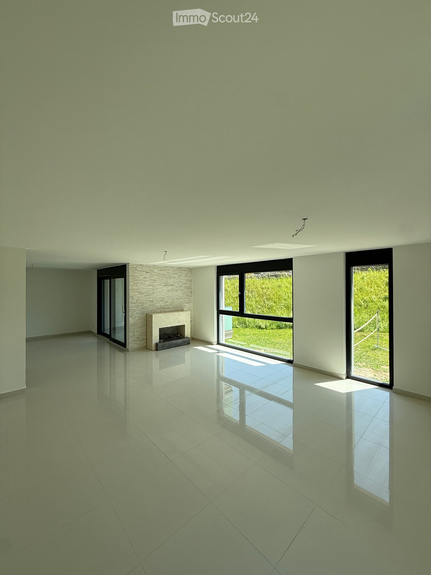 Empty room with light green walls, polished tile floor, fireplace, large windows with view of greenery, and modern ceiling lighting.