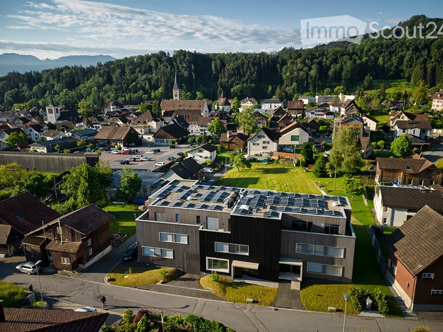 Aerial view of a modern building with solar panels on the roof, surrounded by a small garden and integrated into a village setting with traditional houses and green spaces.