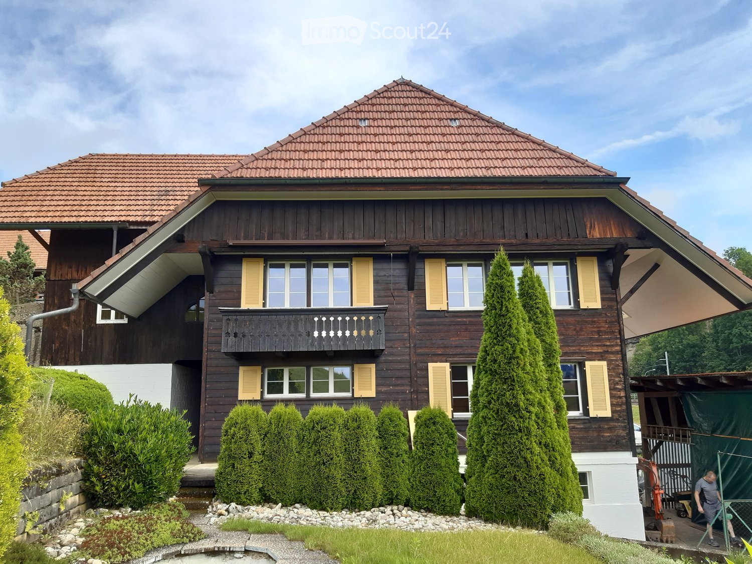Two-story wooden house with a red-tiled roof, multiple windows with shutters, and a balcony on the upper floor. The house is surrounded by well-maintained greenery and shrubs.