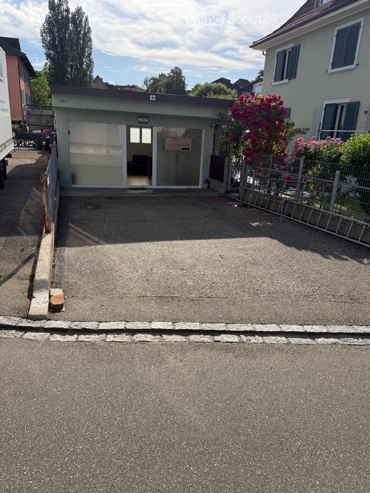 Driveway leading to a small building with a sign reading "ZU VERMIETEN" (For Rent). The building has a modern design with large glass windows and a small seating area visible inside. There is a fence with flowering plants and trees in the background.
